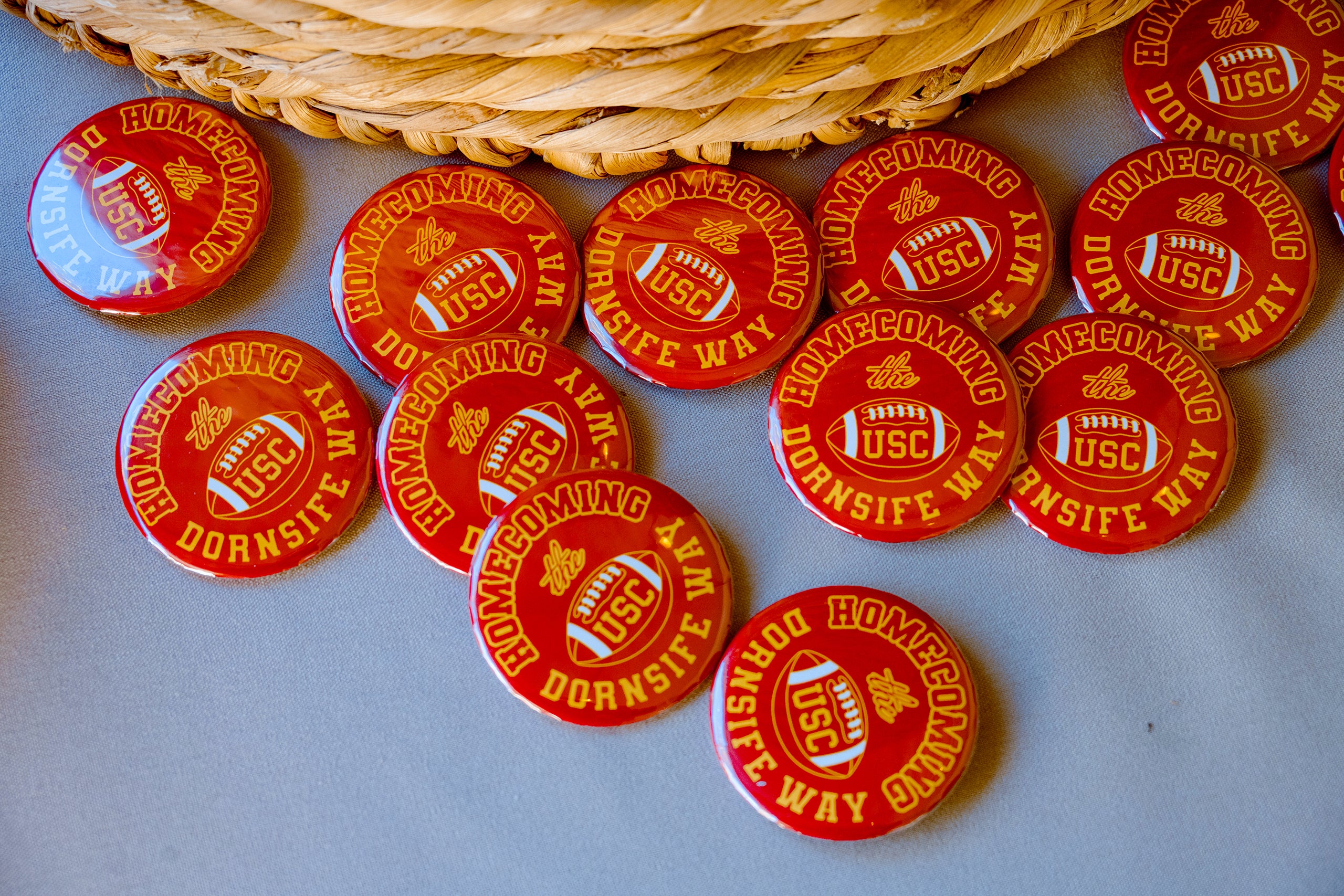 Several red buttons with gold text, reading 'Homecoming the USC Dornsife Way,' are scattered on a table next to a woven basket.