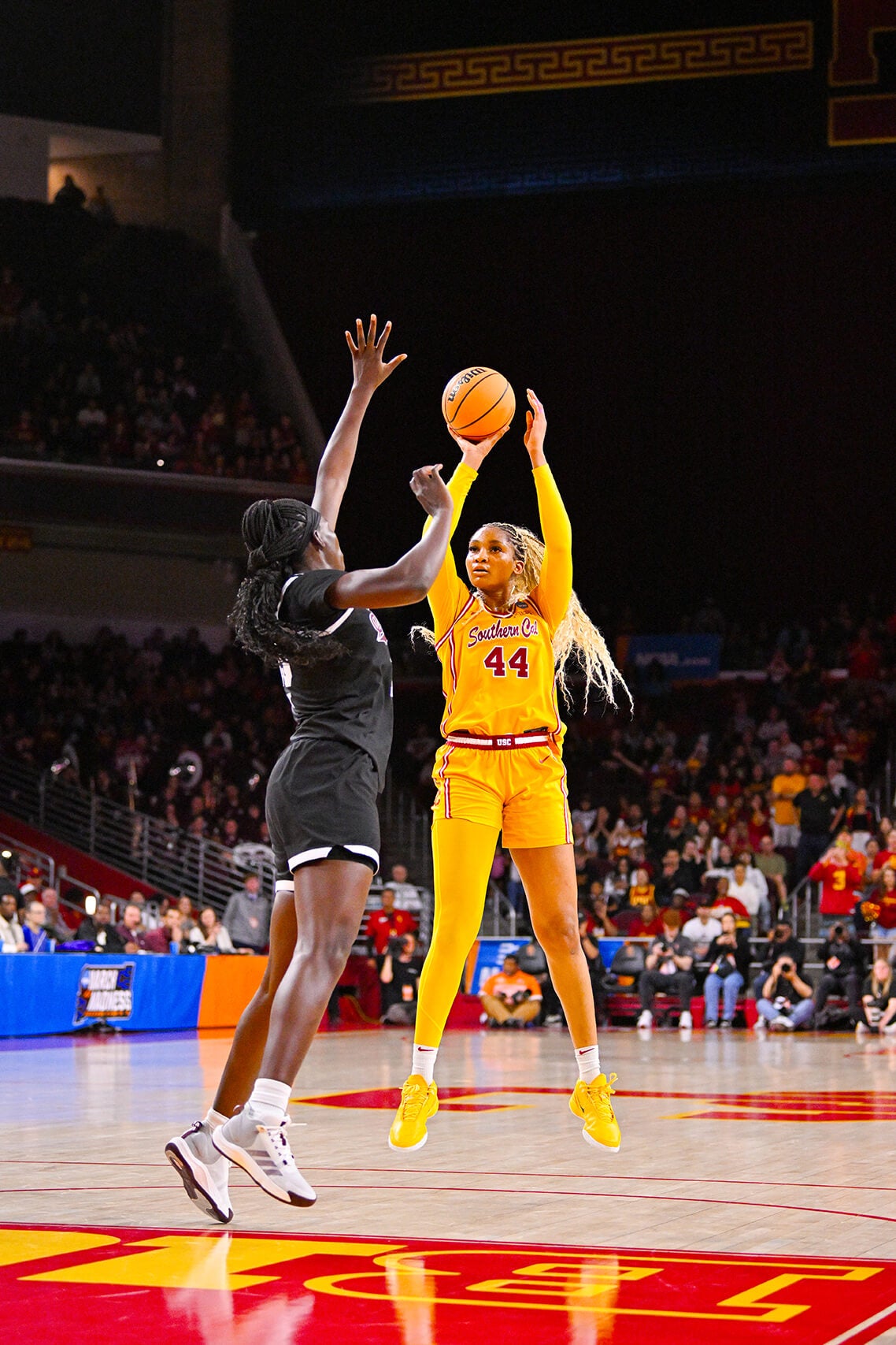 A USC women's basketball player in a yellow uniform shoots over an opponent in black during a game, with a crowd watching in an indoor arena.