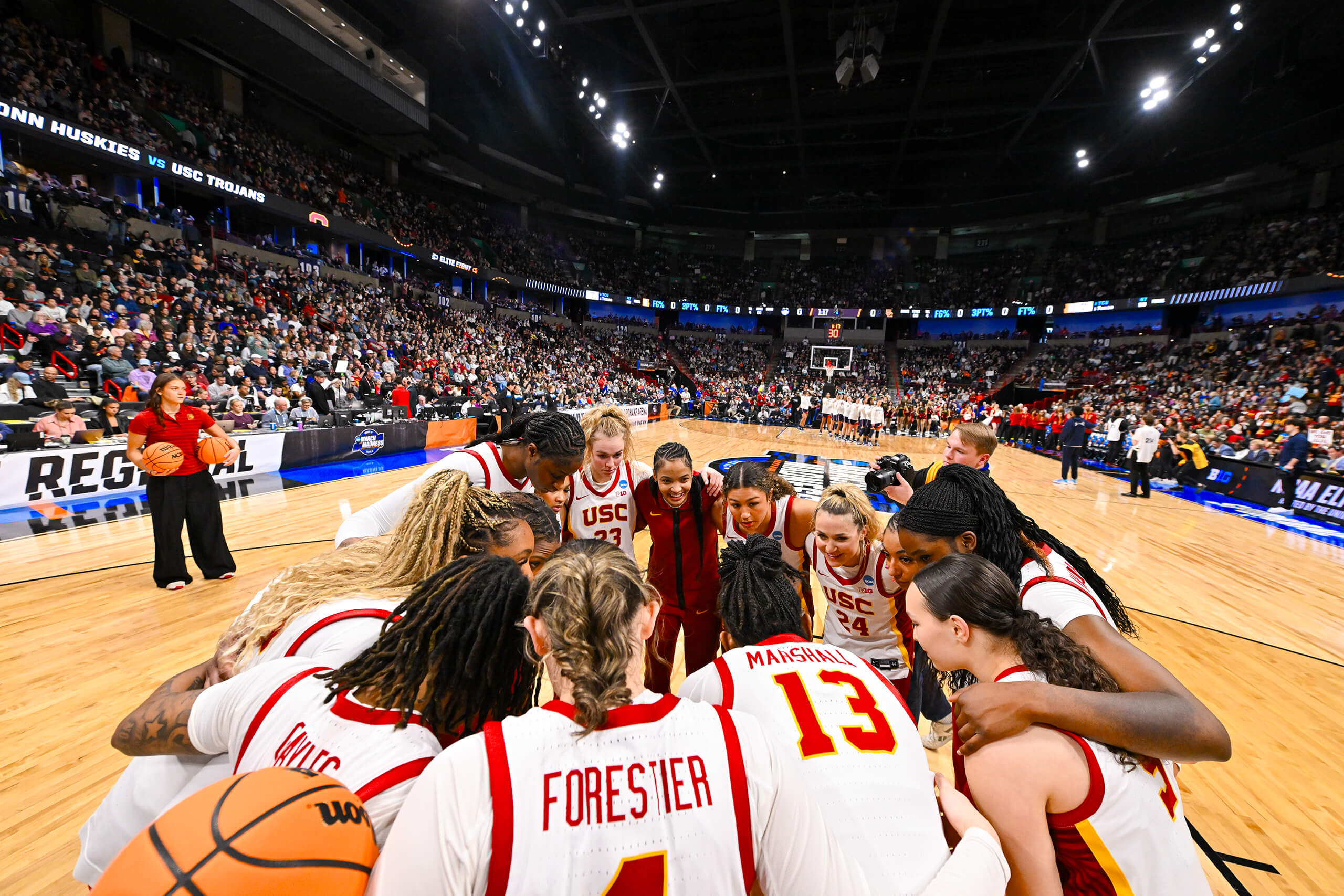 The USC women's basketball team in white jerseys huddles on the court during a game, with a large crowd in the arena and a scoreboard in the background.