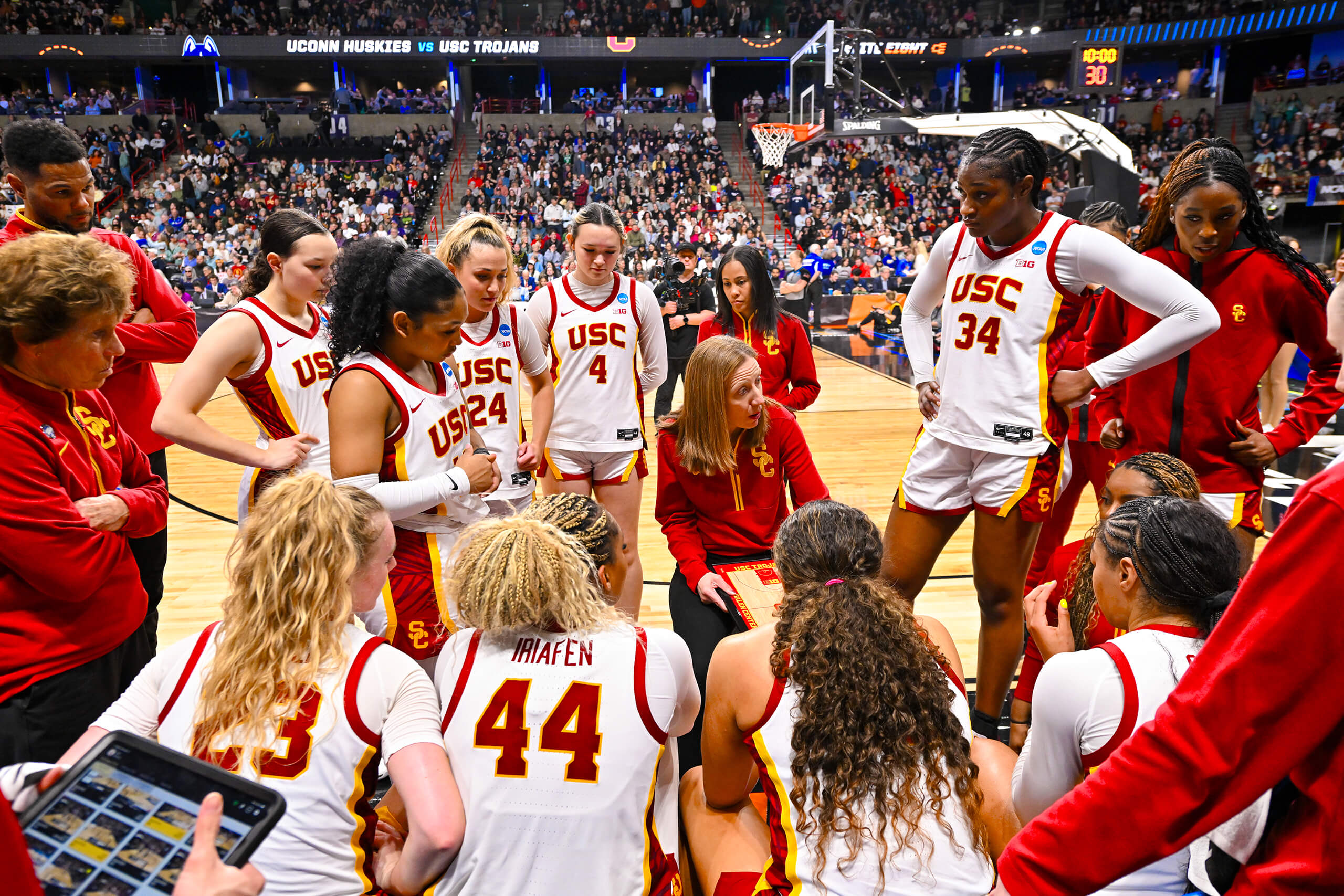 The USC women's basketball team in white jerseys gathers for a timeout during a game, with a coach instructing and a large crowd in the arena.