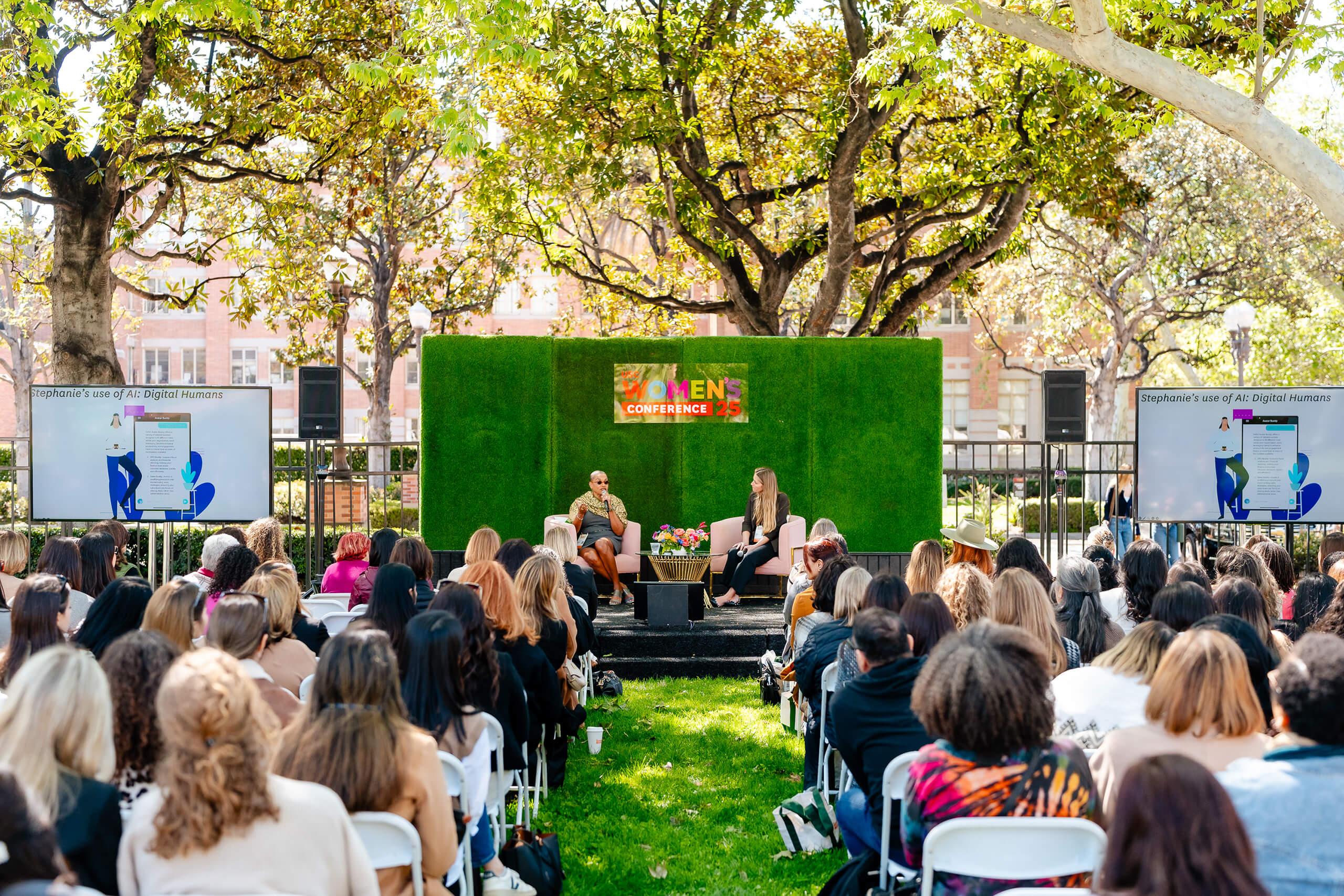 Two women speak at the USC Women's Conference 2025 on a stage with a green backdrop, addressing an audience seated on a grassy area under trees, with screens on either side.