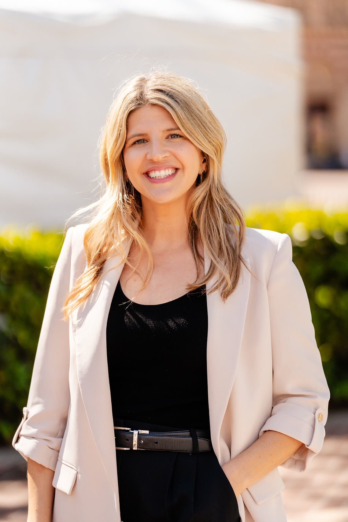 A woman in a light blazer and black outfit stands outdoors at the 2025 USC Women's Conference, with greenery and a brick building in the background.
