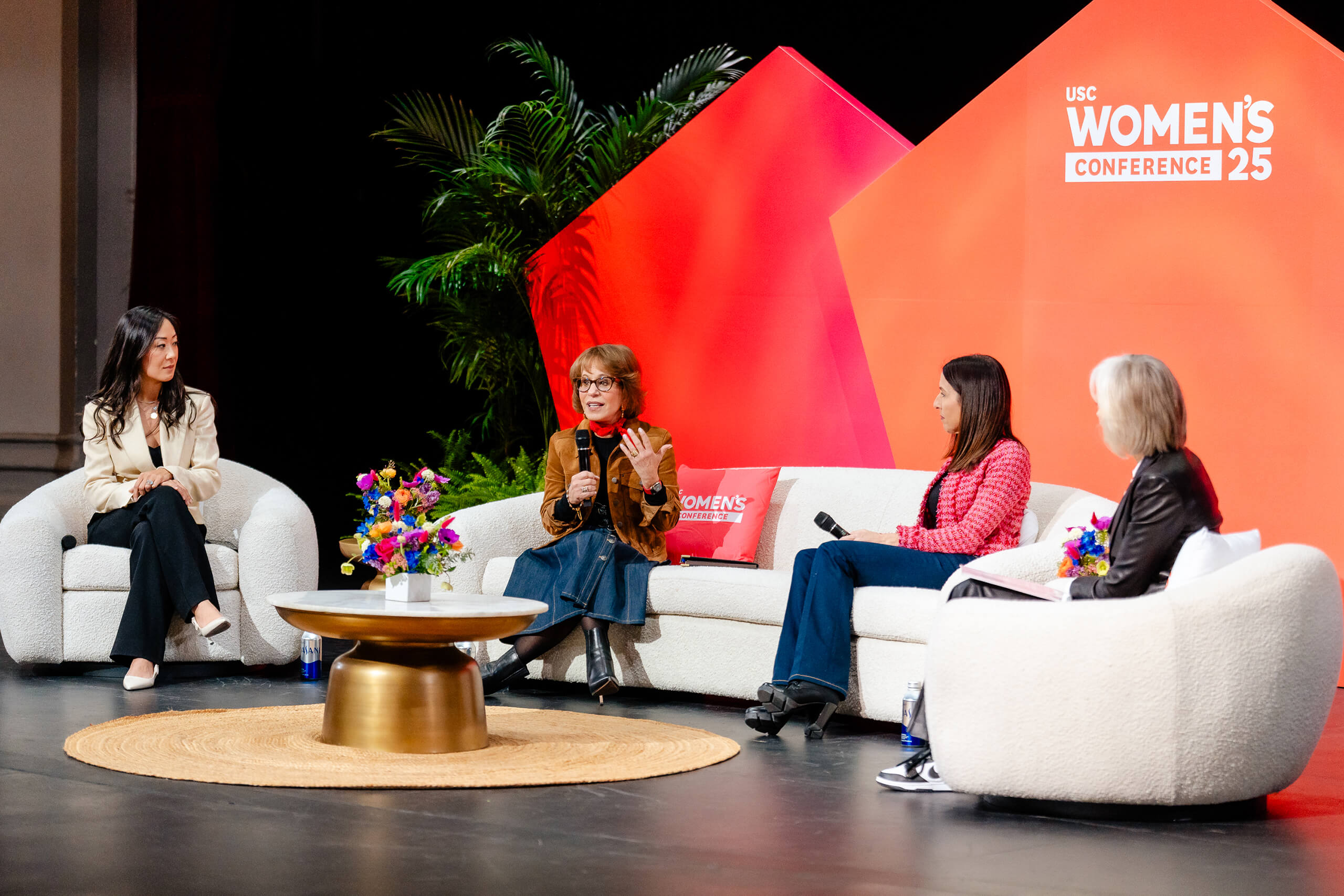 Four women on a stage at the USC Women's Conference 2025, seated on white chairs and a sofa, with a floral centerpiece and a red backdrop, engaging in a panel discussion.