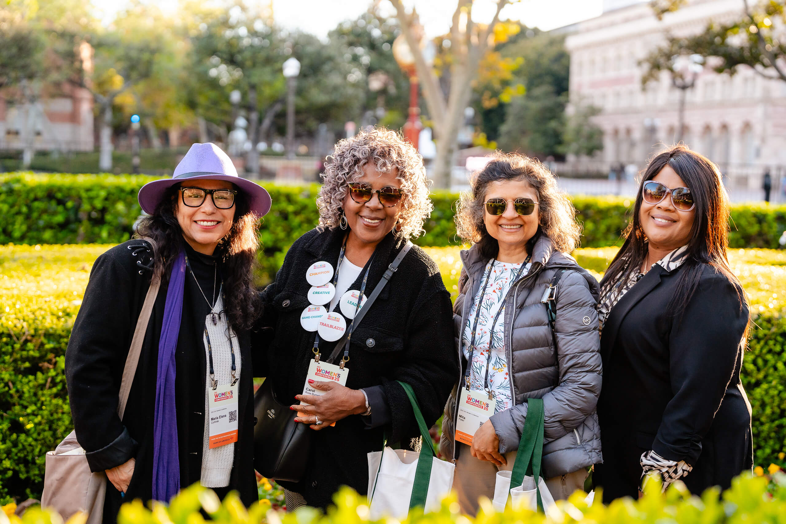 Four women with conference badges pose outdoors at the 2025 USC Women's Conference, smiling in front of greenery and a brick building, holding tote bags.
