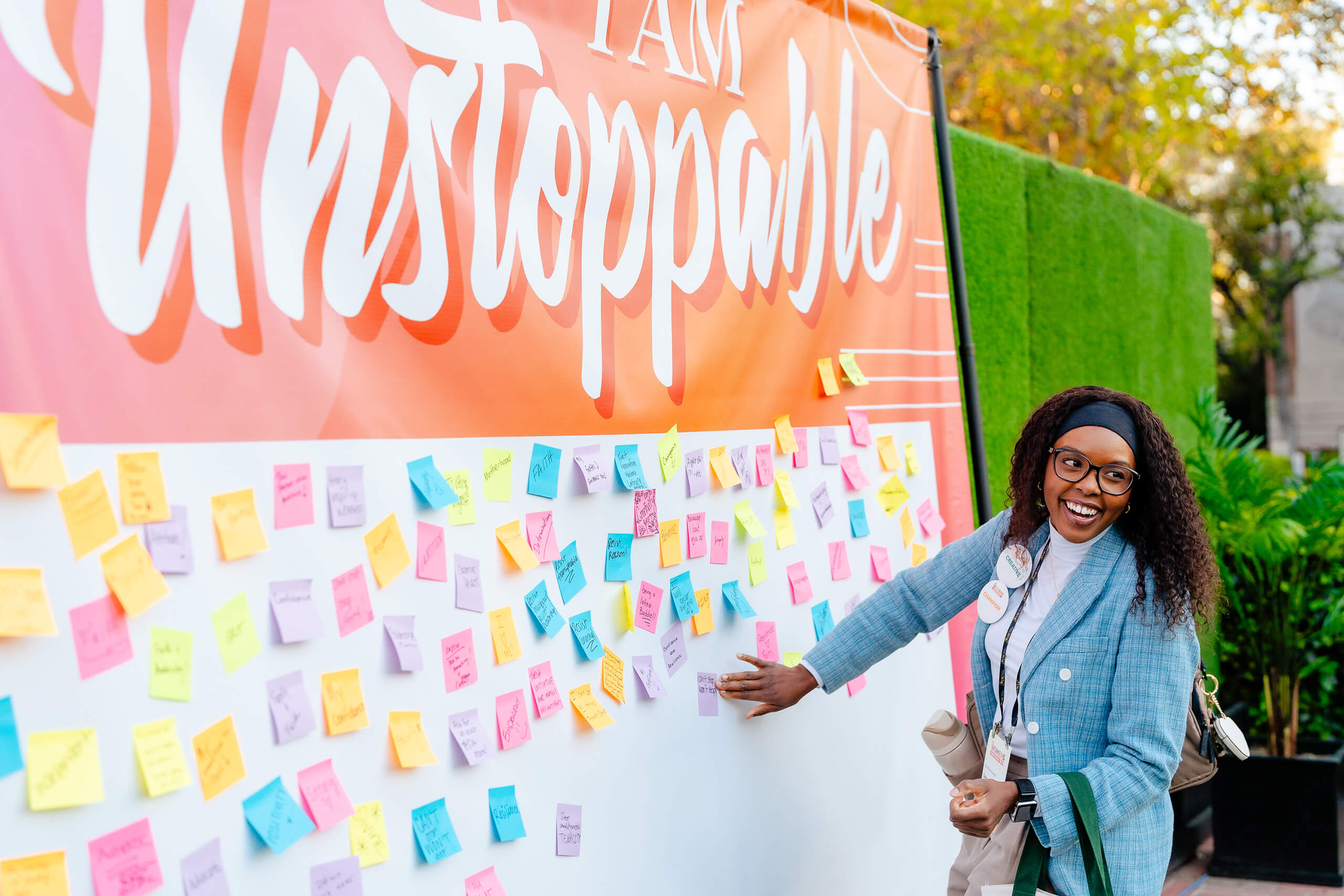 A woman in a blue blazer adds a sticky note to a board with many colorful notes, in front of a 'I AM UNSTOPPABLE' banner at the 2025 USC Women's Conference.