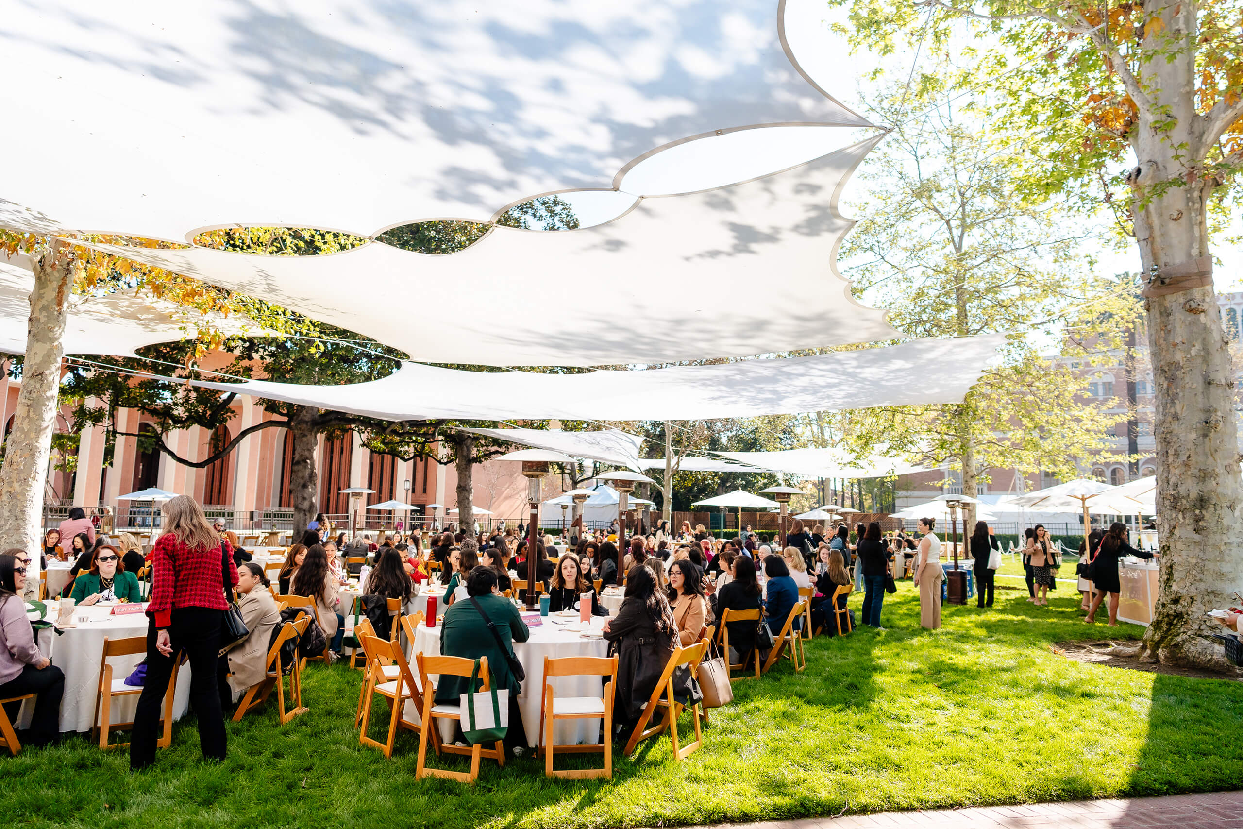 A large group of women sit at tables under white canopy shades on a grassy area, engaging in conversation at the 2025 USC Women's Conference, with trees and buildings around.