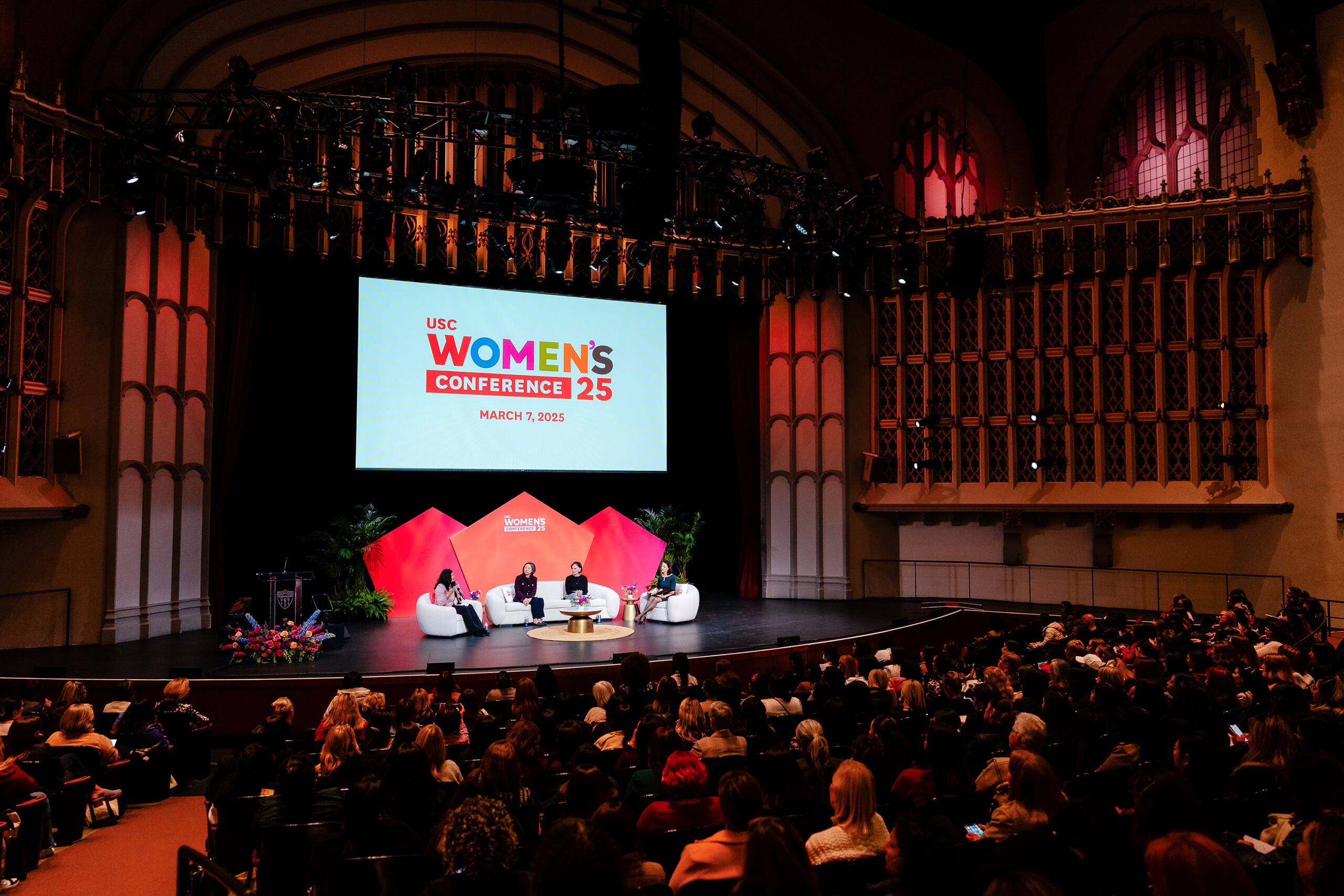 A panel of four women speak at the USC Women's Conference 2025 on March 7, in the Bovard Auditorium with a large audience, a screen displaying the event details, and floral decor.