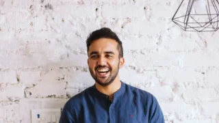 Ajai Thandi smiling against a white brick wall, wearing a blue shirt, under a geometric light.