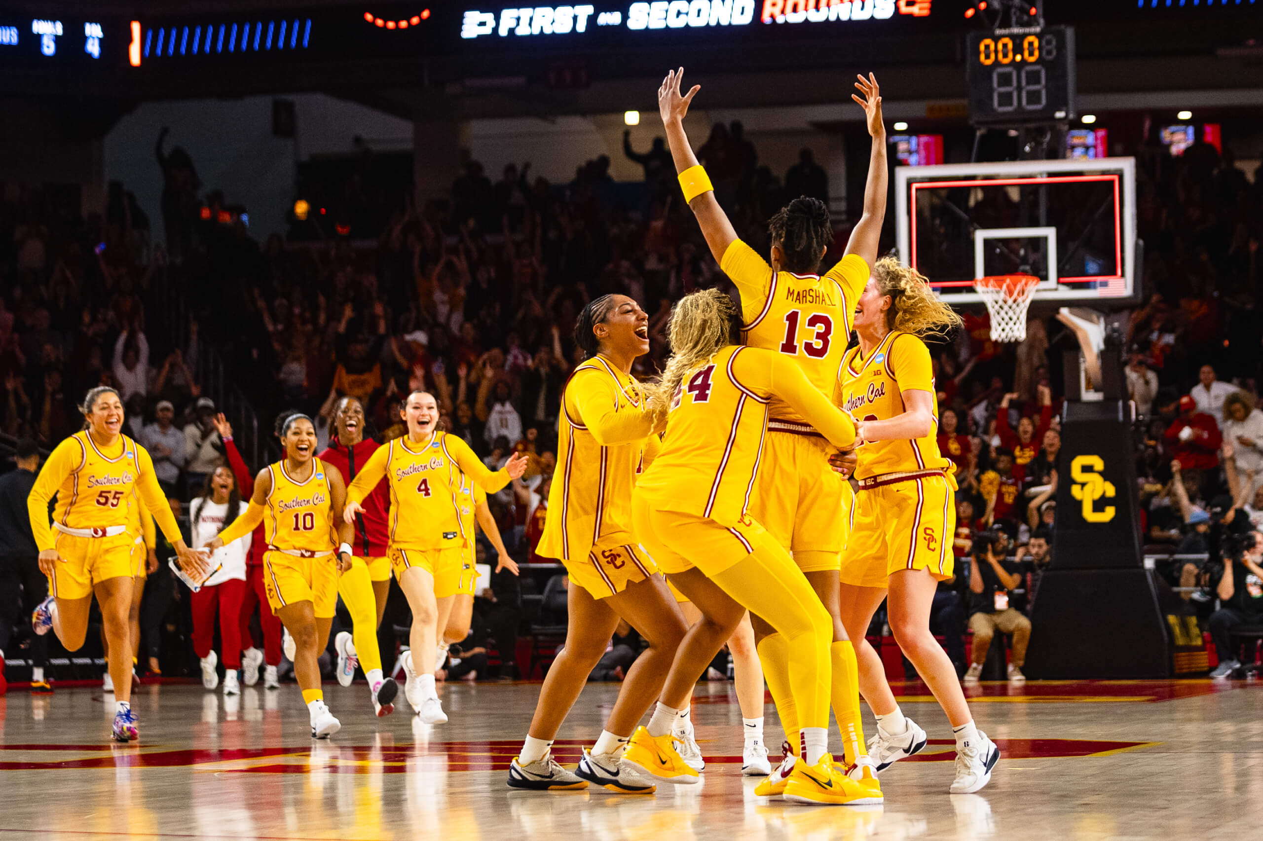 USC women's basketball team in yellow uniforms celebrates on the court between quarters, with a crowd cheering in the arena and the scoreboard showing the break.