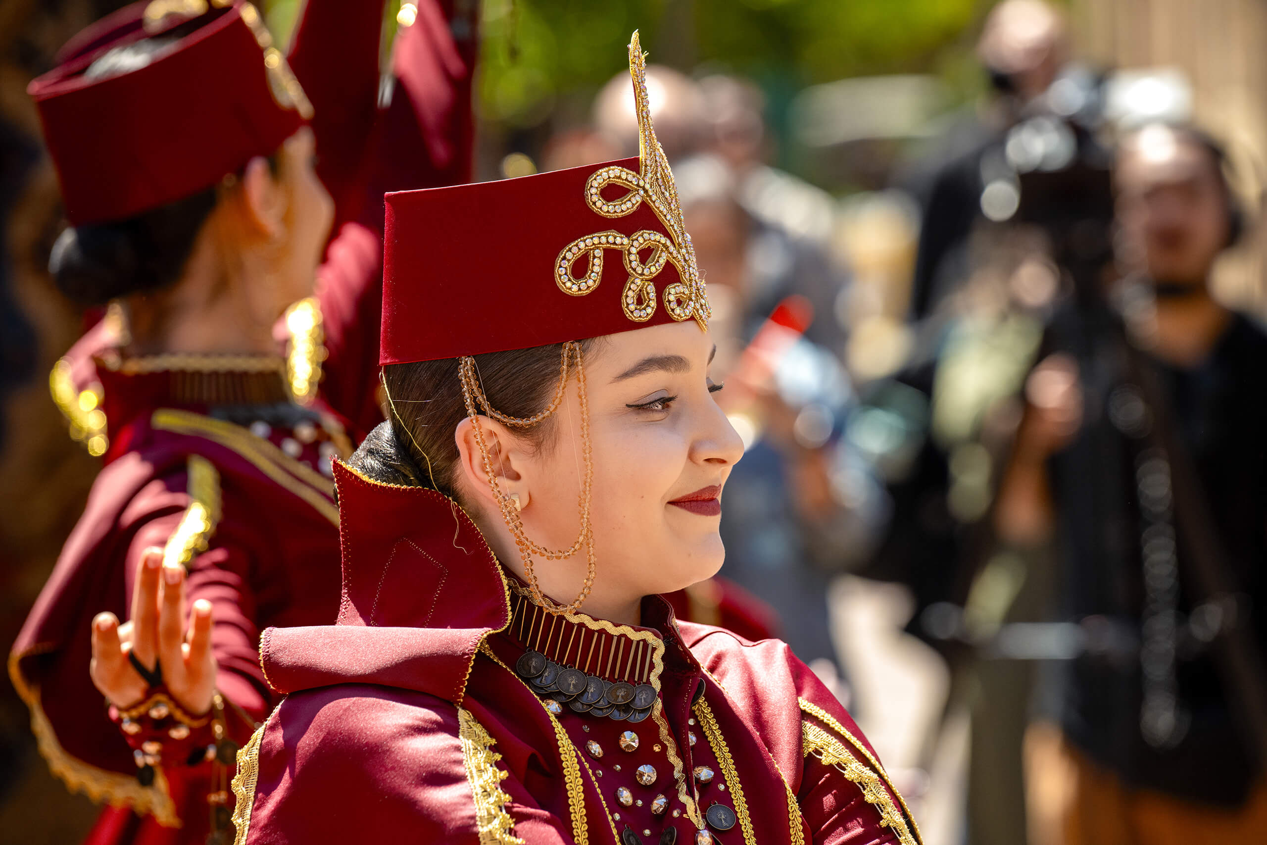 A woman in a red traditional dress with gold embroidery and a matching hat, adorned with jewelry, performs a dance in a crowd, others in similar attire visible.