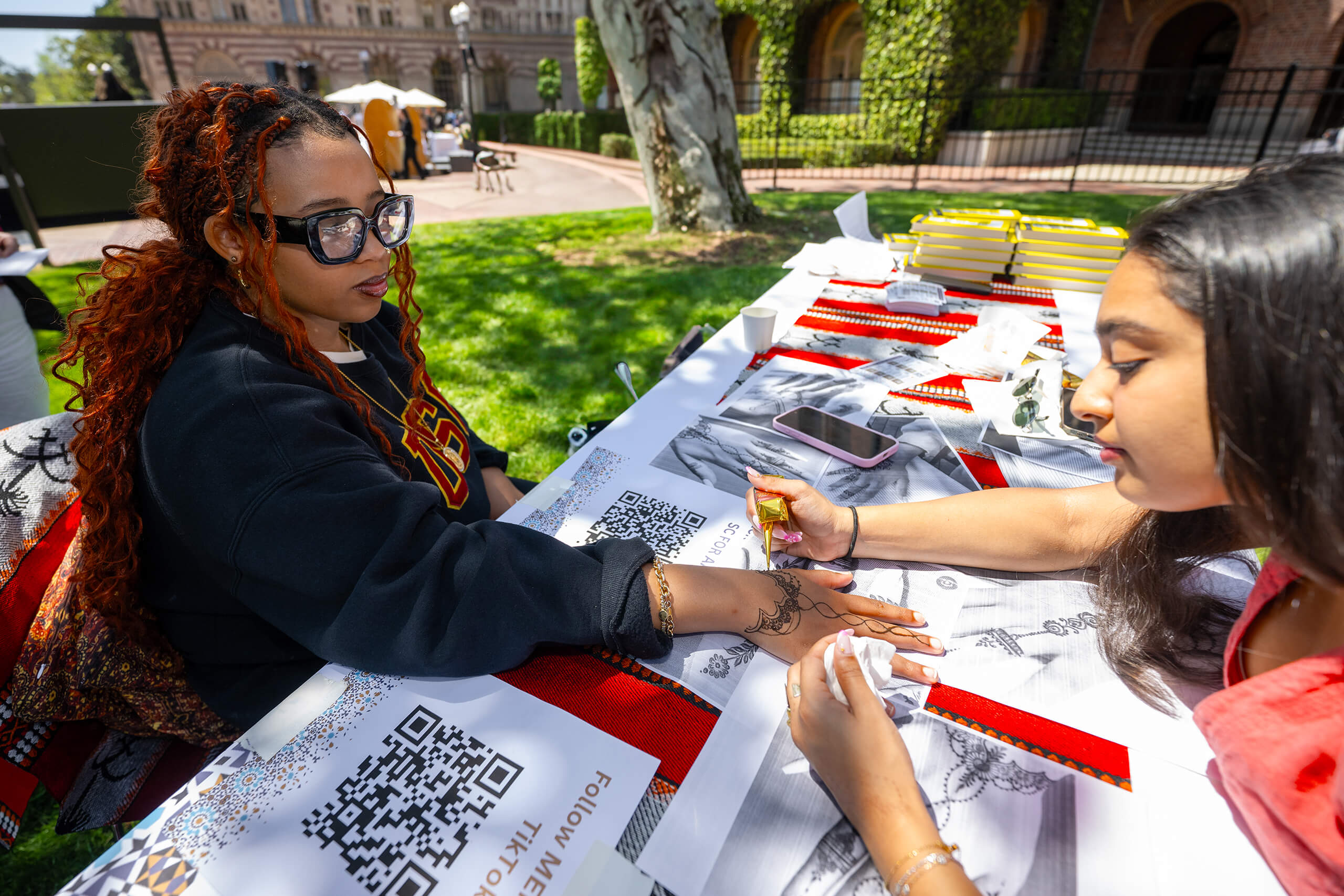 Two women at an outdoor table at the USC Campus. One applies henna to the other's hand.