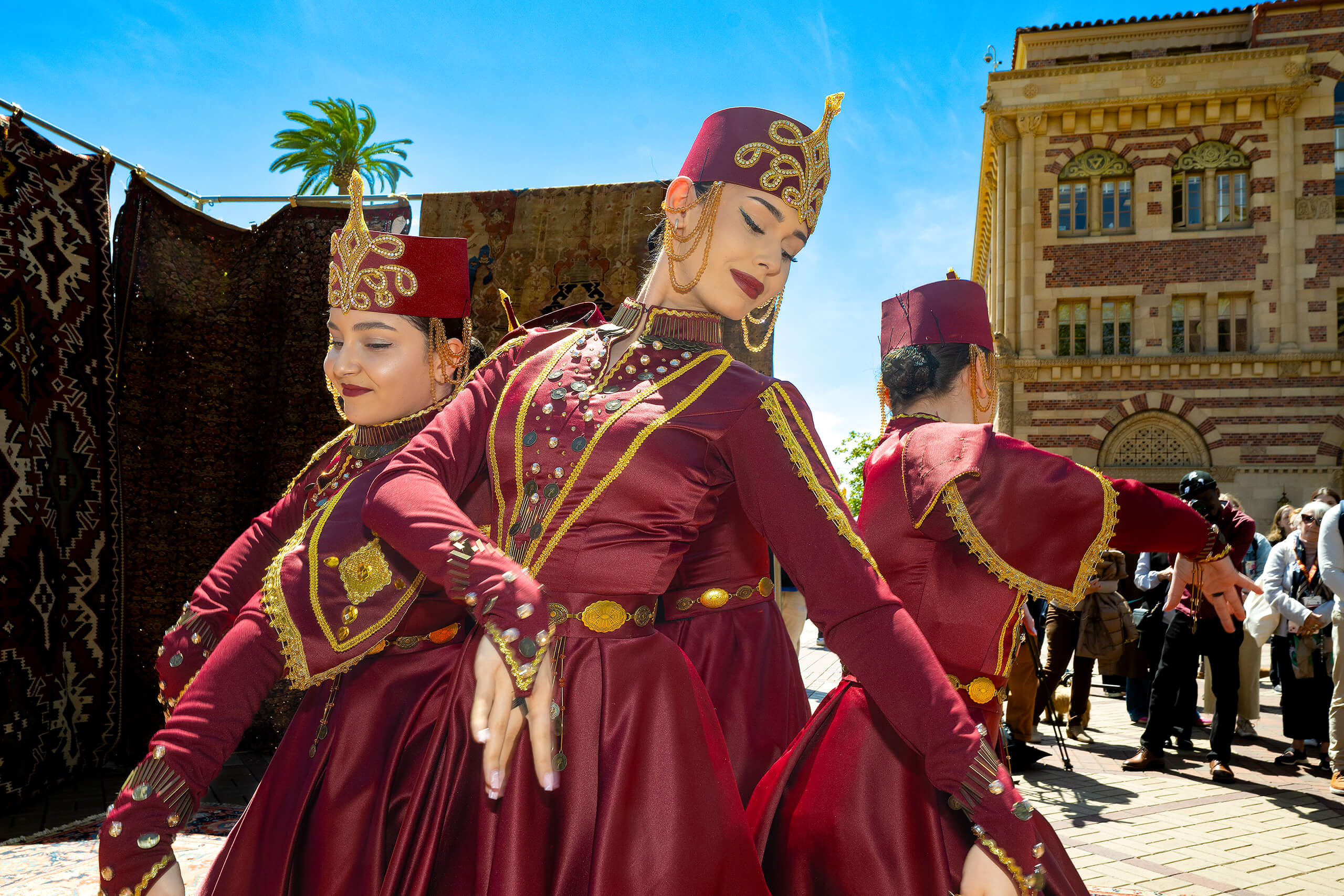 Three women in red traditional dresses with gold embroidery dance outdoors, with patterned rugs, a palm tree, and a brick building in the background, crowd watching.