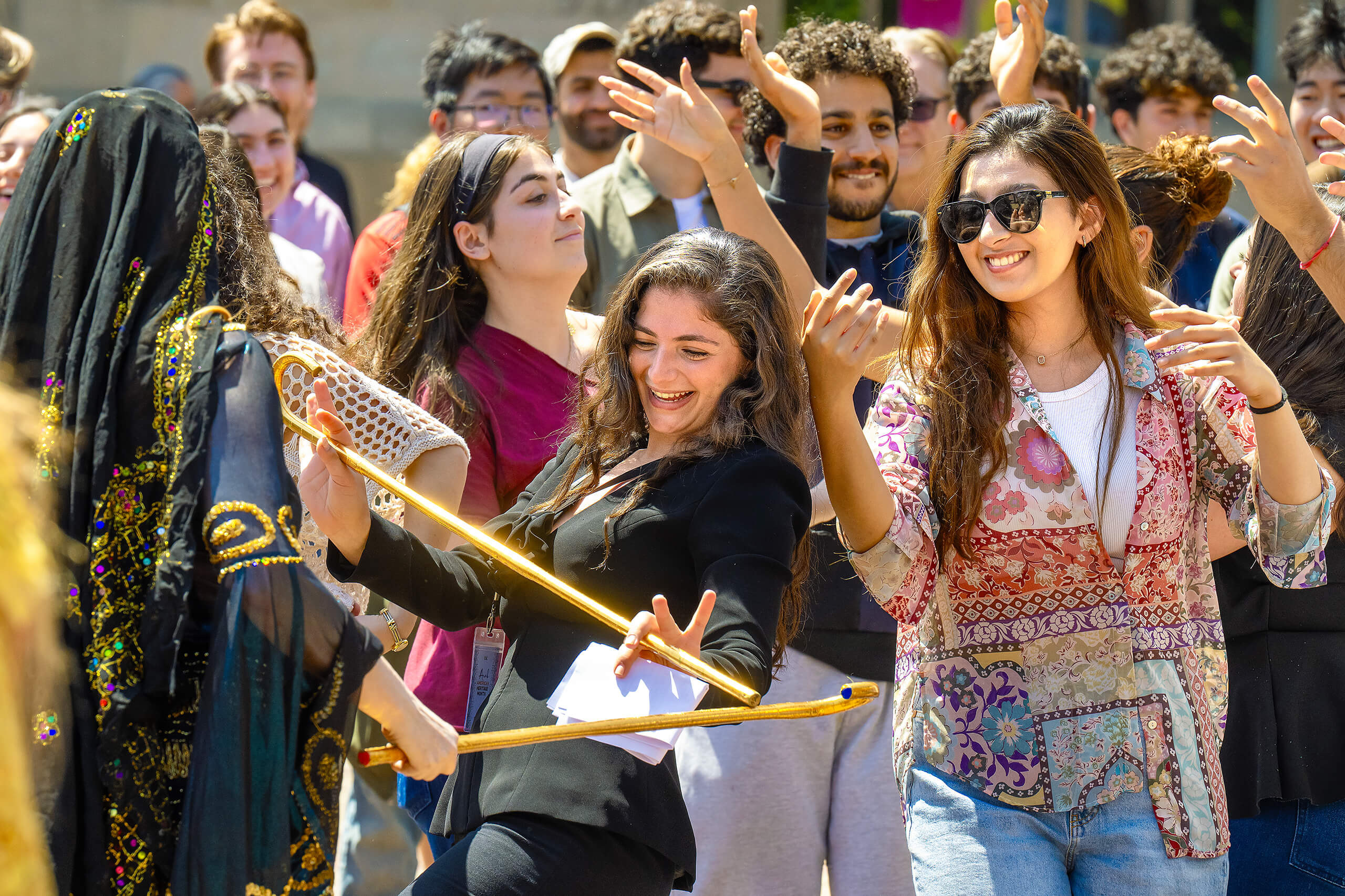 A group of people, some in traditional attire, dance and clap hands in a crowd, one holding a golden staff, others in casual clothing, at an outdoor event.