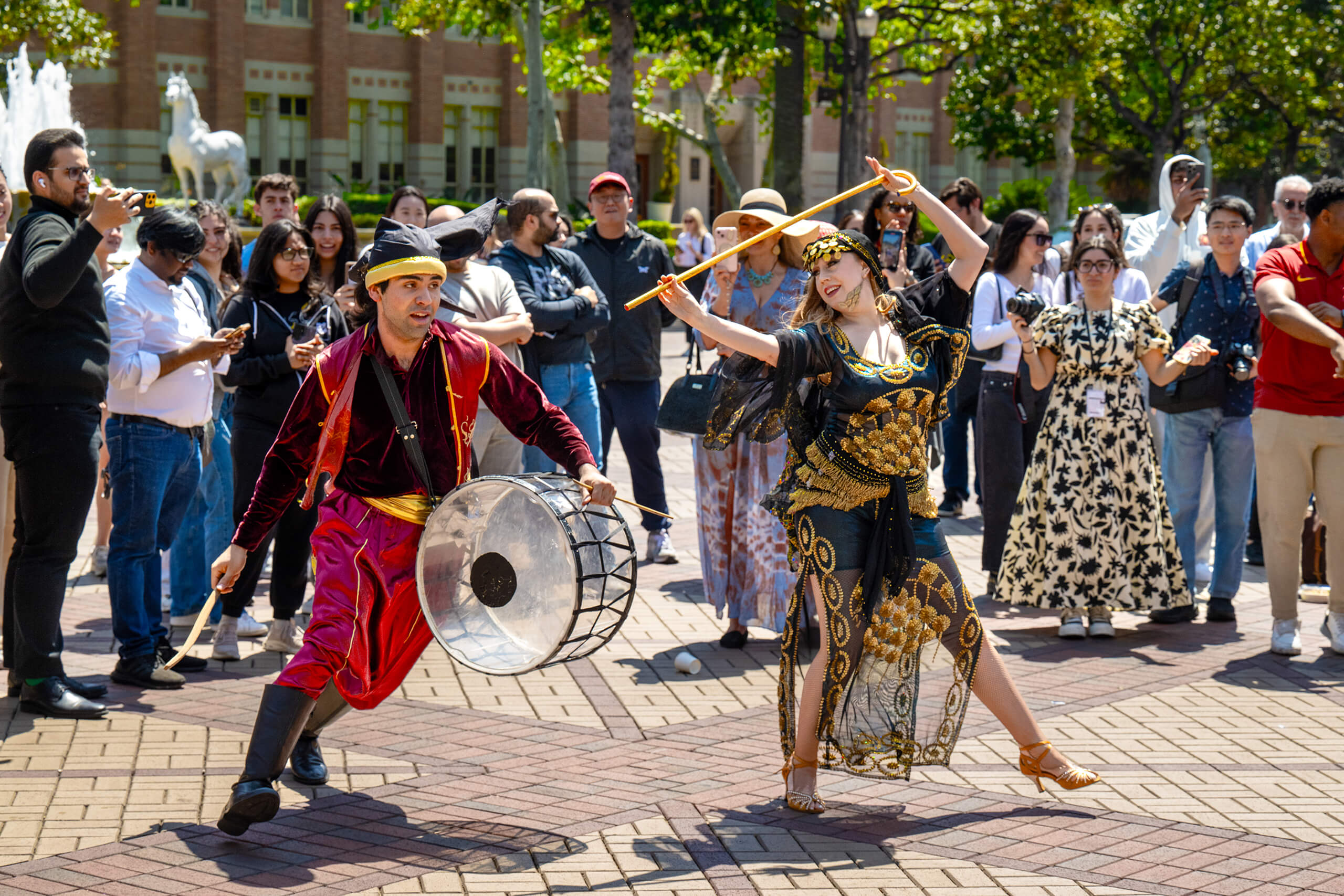 A man in red traditional attire plays a drum while a woman in a black and gold outfit dances with a staff, a crowd watches in an outdoor setting.
