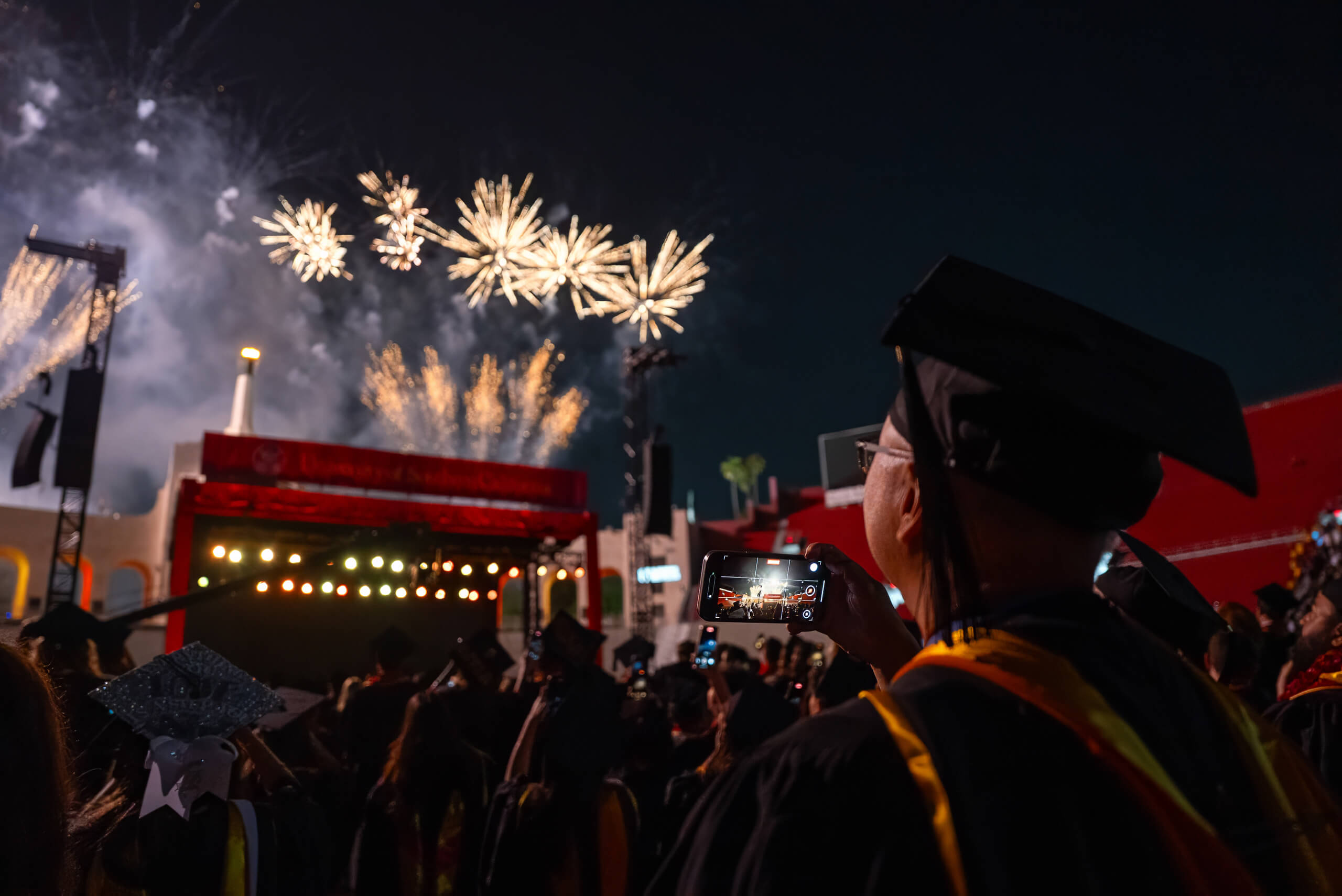 A graduate in regalia films fireworks lighting up the night sky during the 2025 USC commencement ceremony at the Los Angeles Coliseum.