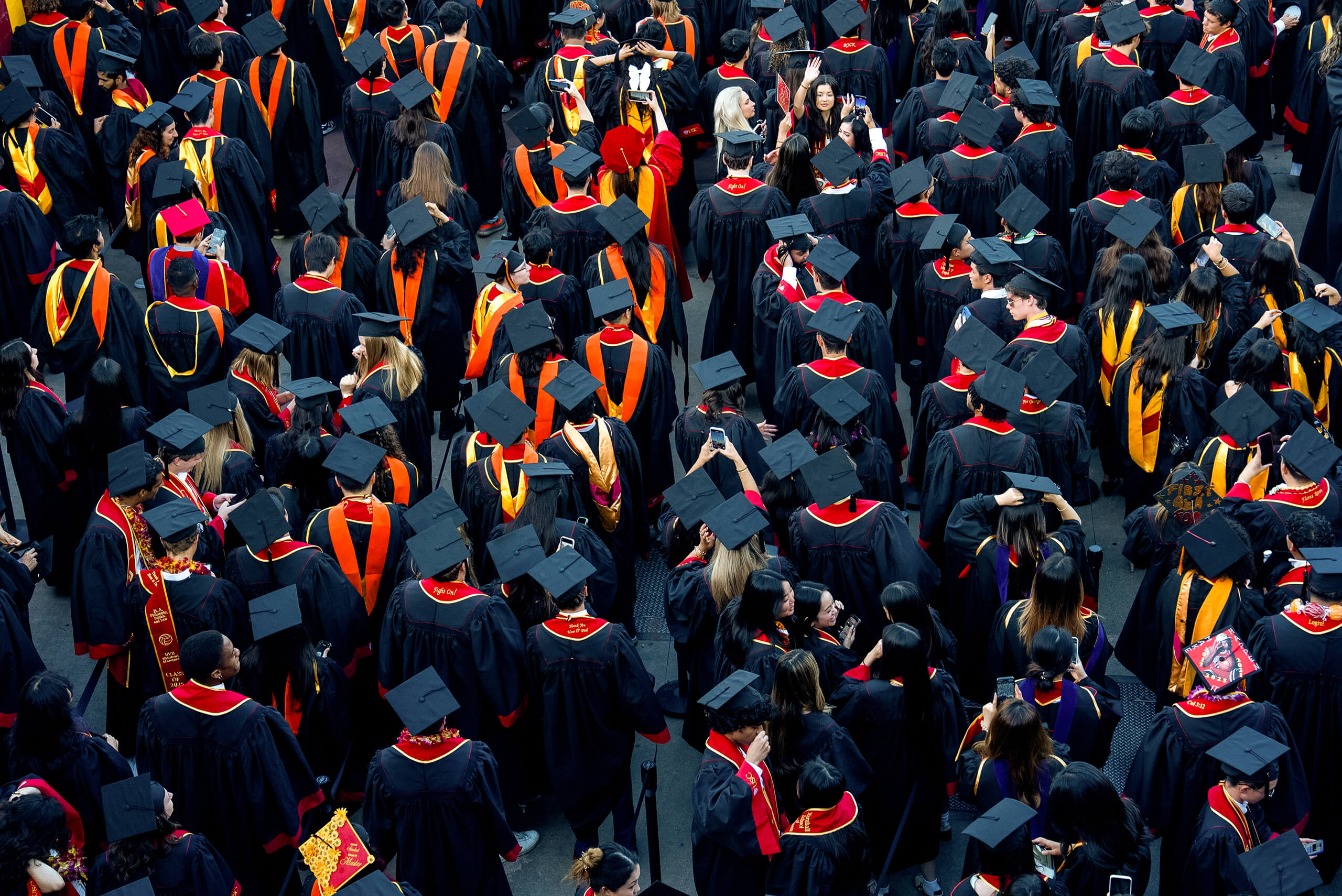 A large crowd of USC graduates in black gowns with red and gold accents gathers, taking photos and celebrating at the 2025 commencement ceremony.