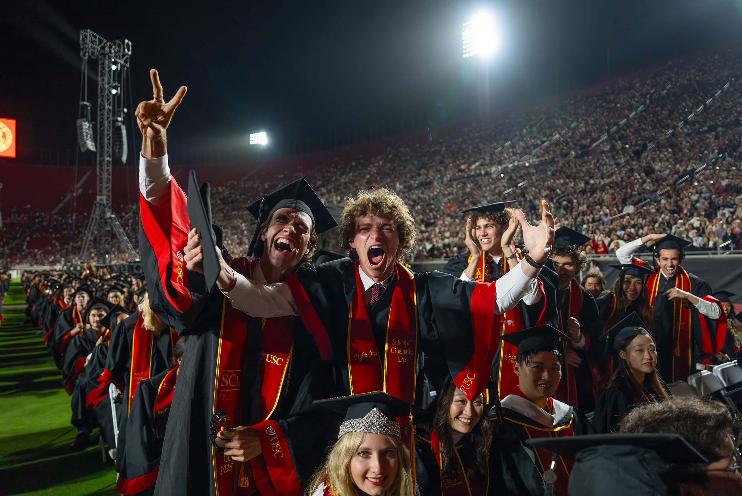 Excited USC graduates in red and gold regalia from the Class of 2025 cheer and raise hands at a ceremony in the Los Angeles Coliseum, with a large crowd in the background.