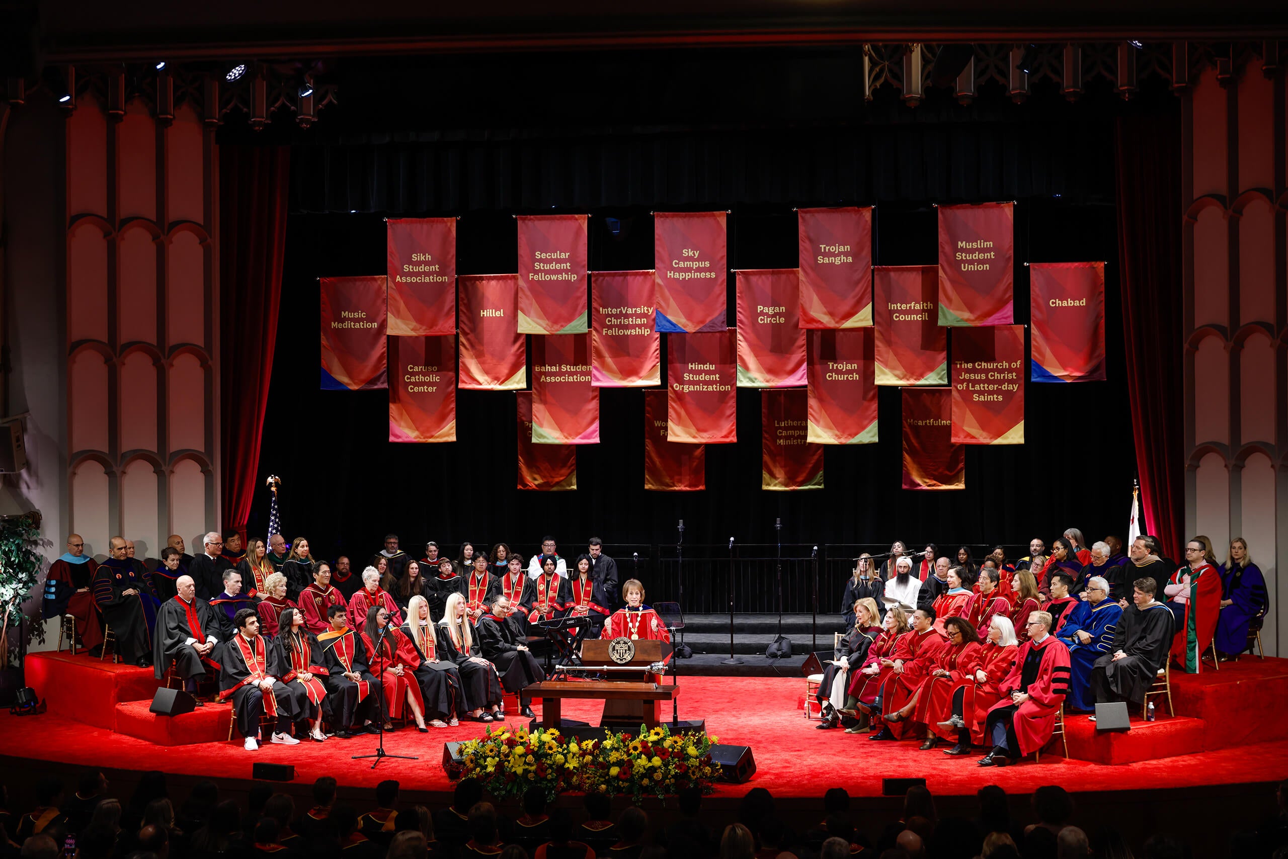 Faculty in regalia sit on a red stage with banners of student organizations at the 2025 USC Baccalaureate in Bovard Auditorium.