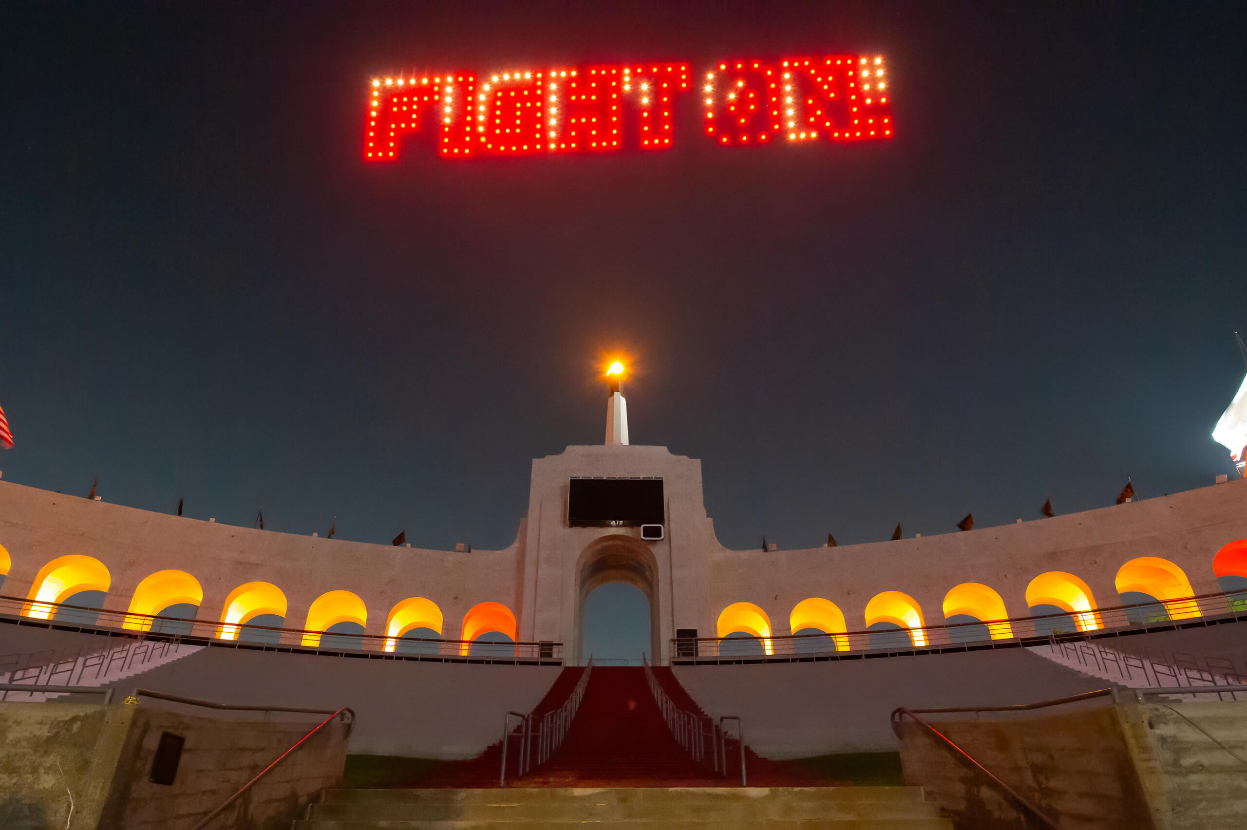 The Los Angeles Coliseum entrance glows with warm lights, a torch atop, and 'FIGHT ON' formed by drones above, celebrating the 2025 USC commencement at night.