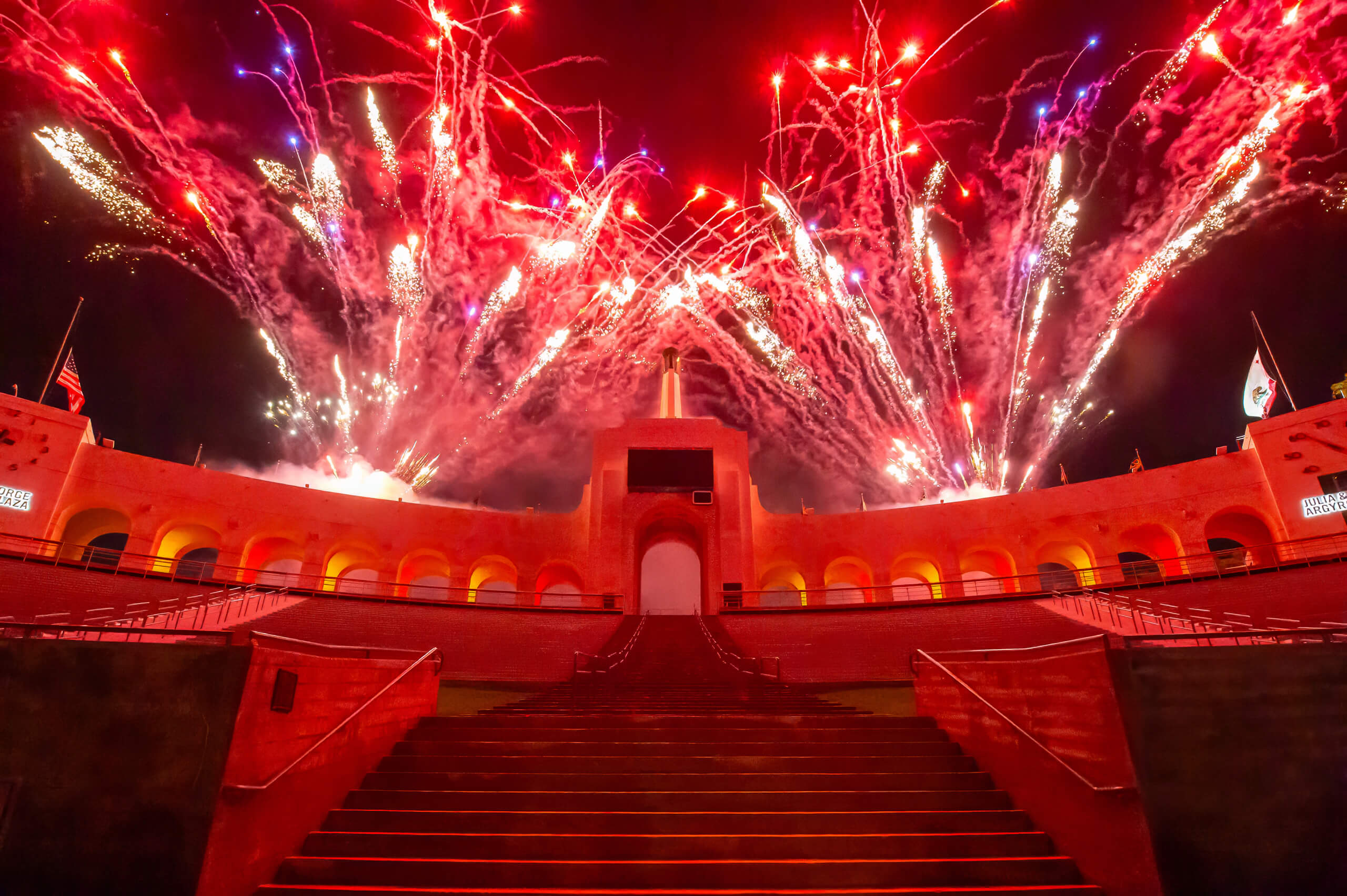 Fireworks light up the night sky over the LA Coliseum during the USC 2025 Commencement Ceremony and red-lit arches in the foreground.