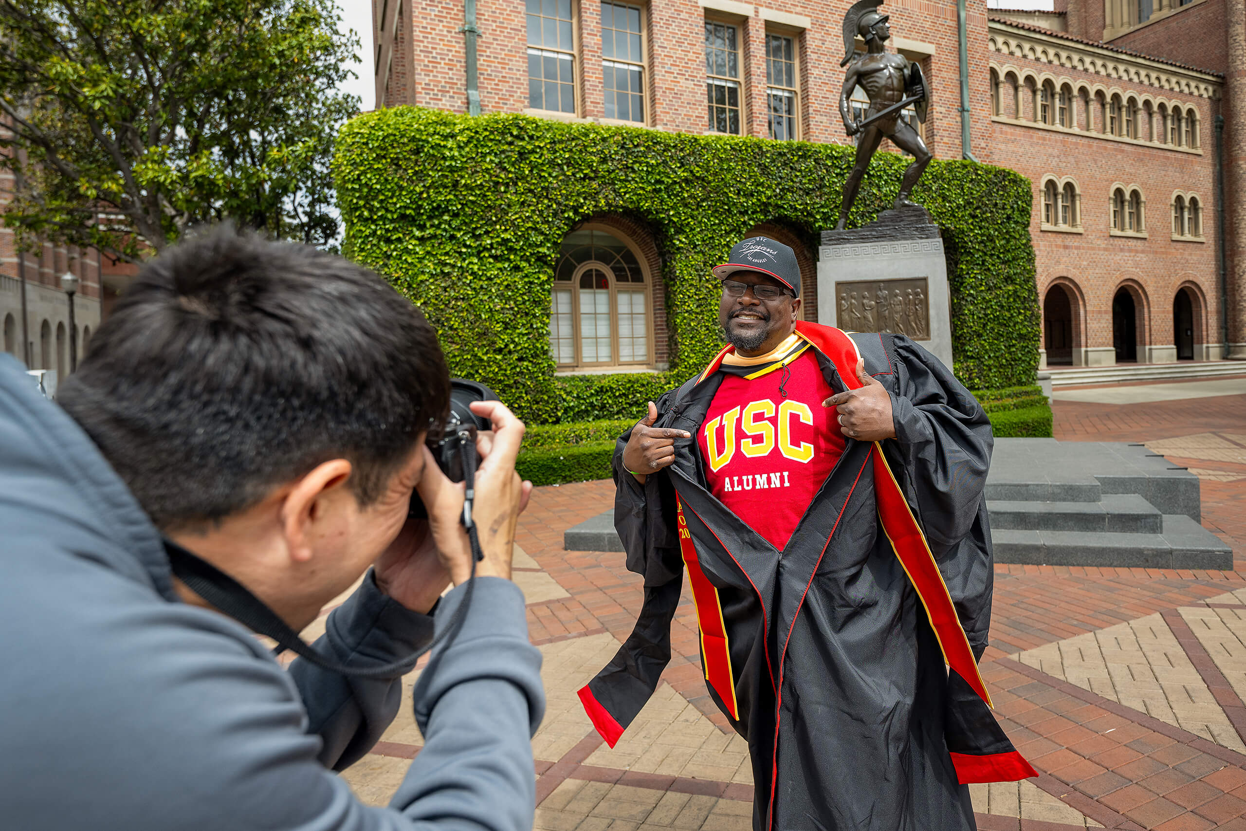 A USC alumnus in red and gold regalia poses, revealing a USC Alumni shirt, while being photographed on campus.