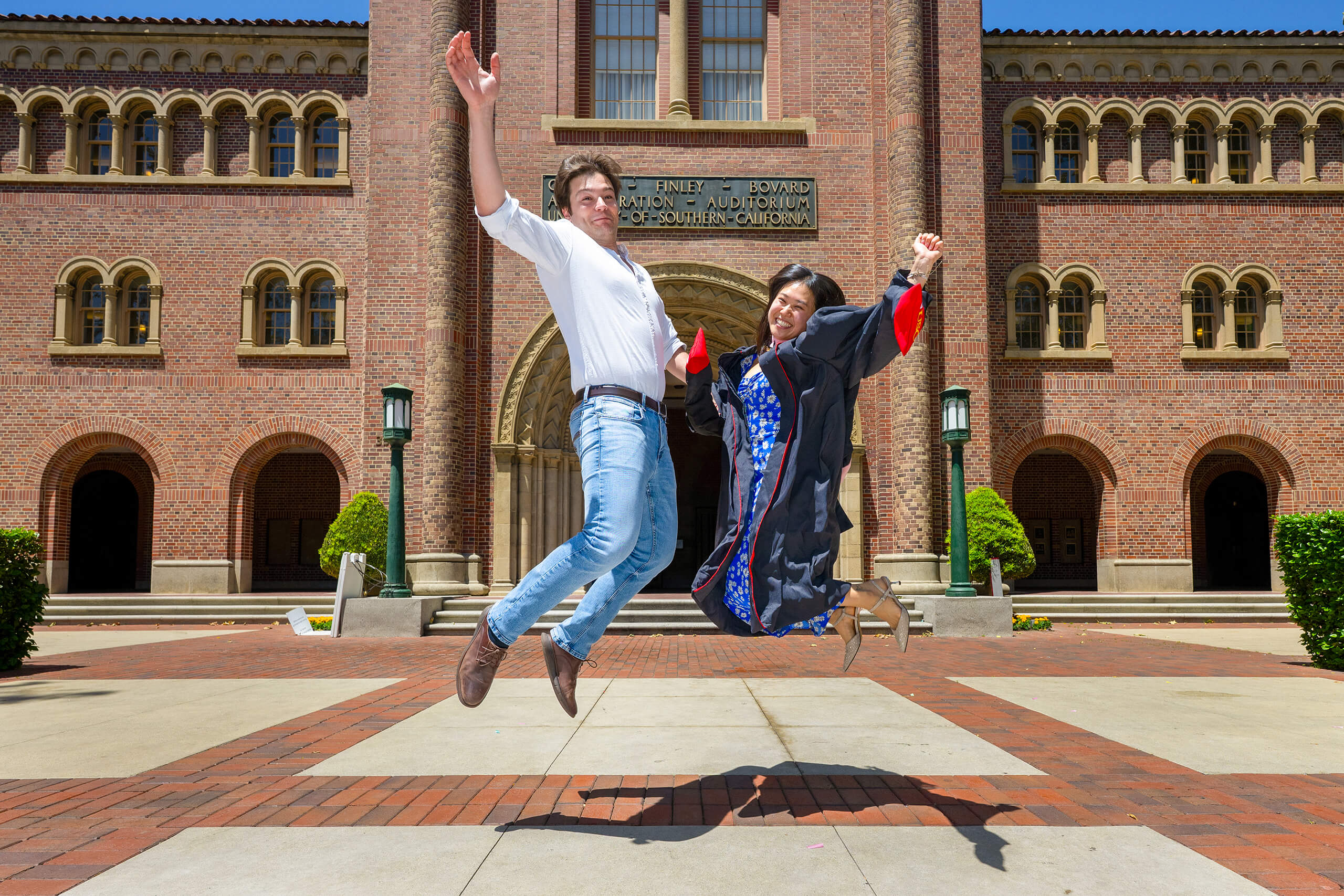 A graduate in USC regalia and a friend leap joyfully in front of Bovard Auditorium during the 2025 commencement celebration.