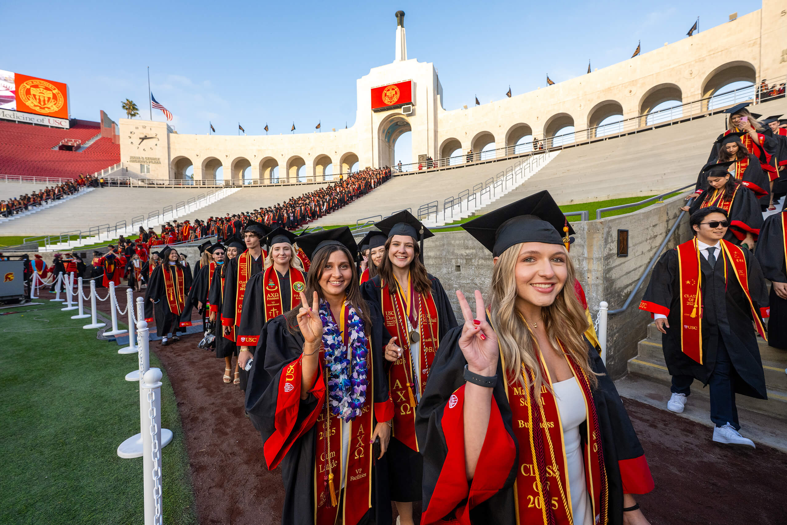 USC graduates in red and gold regalia from the Class of 2025 smile and make the fight on hand gesture while entering the Los Angeles Coliseum for commencement.