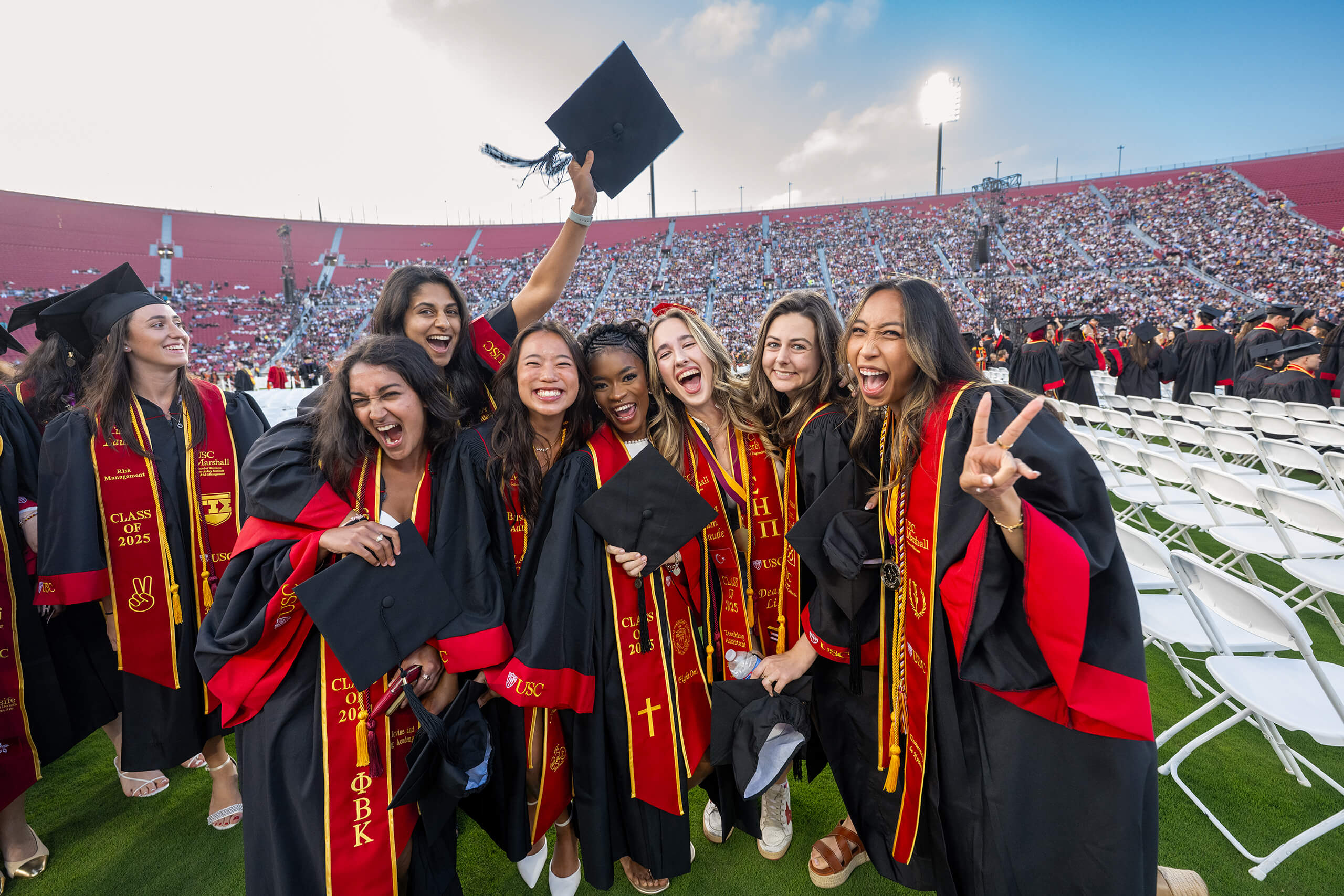 Joyful USC graduates in red and gold regalia from the Class of 2025 celebrate with fight on hand gestures at the Los Angeles Coliseum commencement.