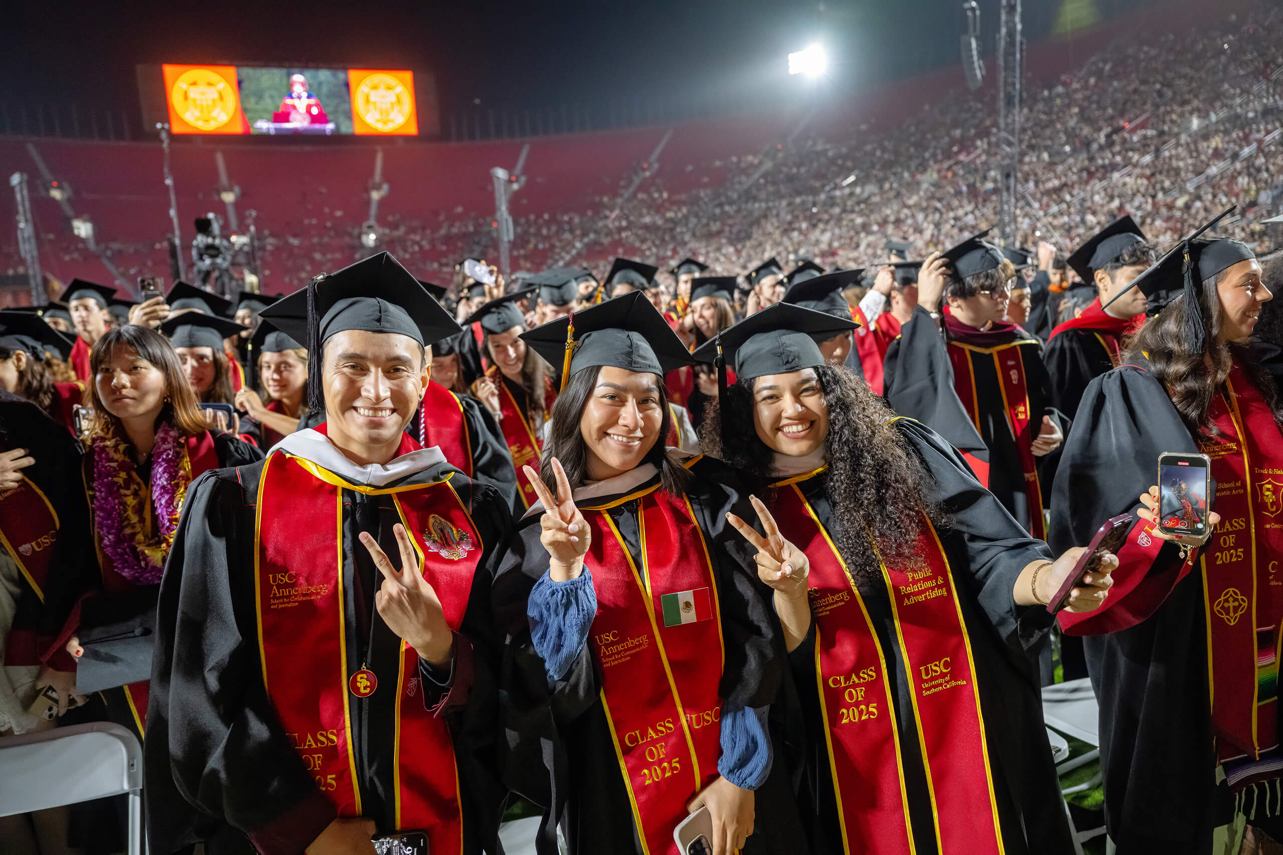 Three USC graduates in red and gold regalia from the Class of 2025 smile and make the fight on hand gesture at a ceremony in the Los Angeles Coliseum, with a large crowd in the background.