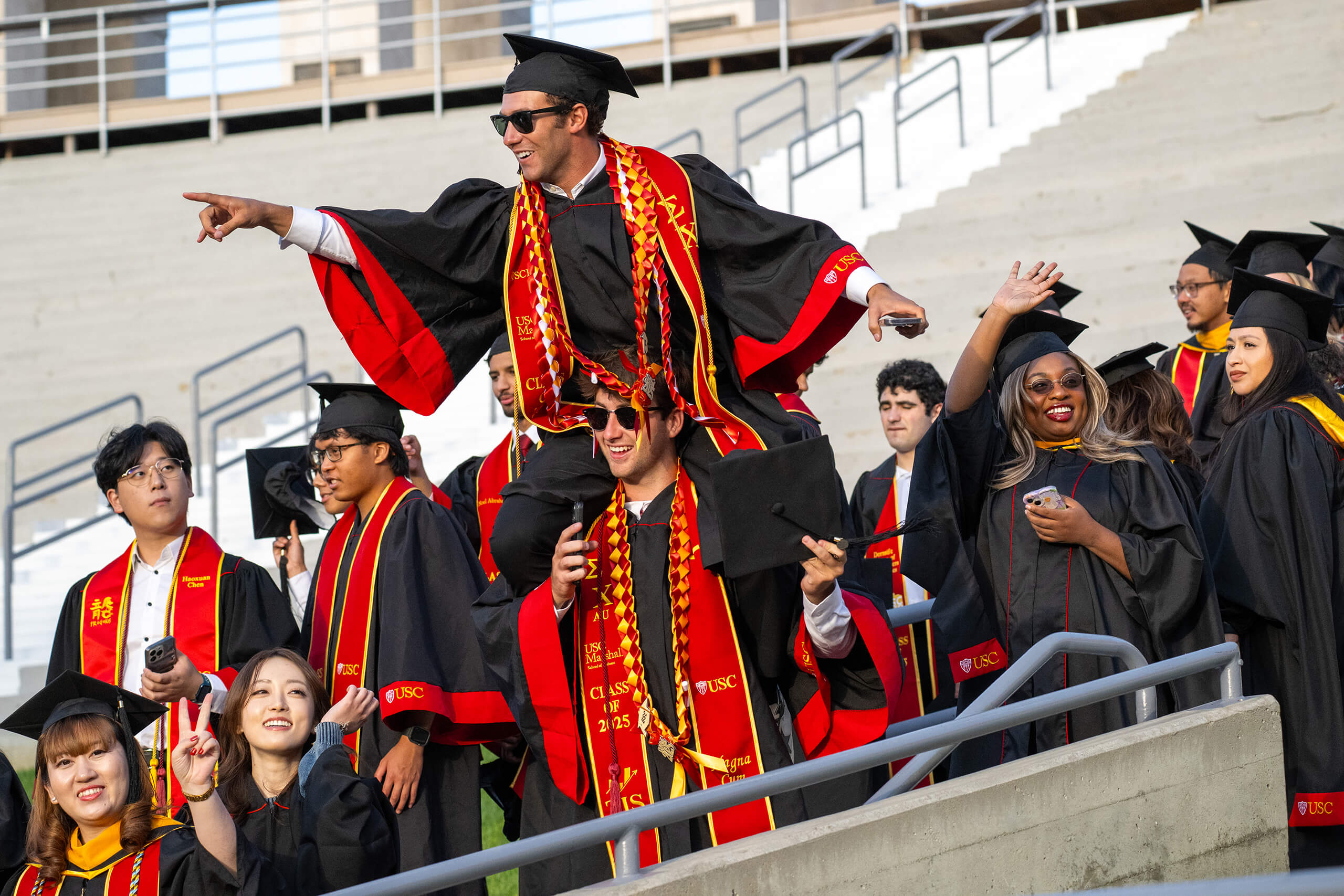 Excited USC graduates in red and gold regalia from the Class of 2025 cheer and make fight on gestures at the Los Angeles Coliseum commencement.