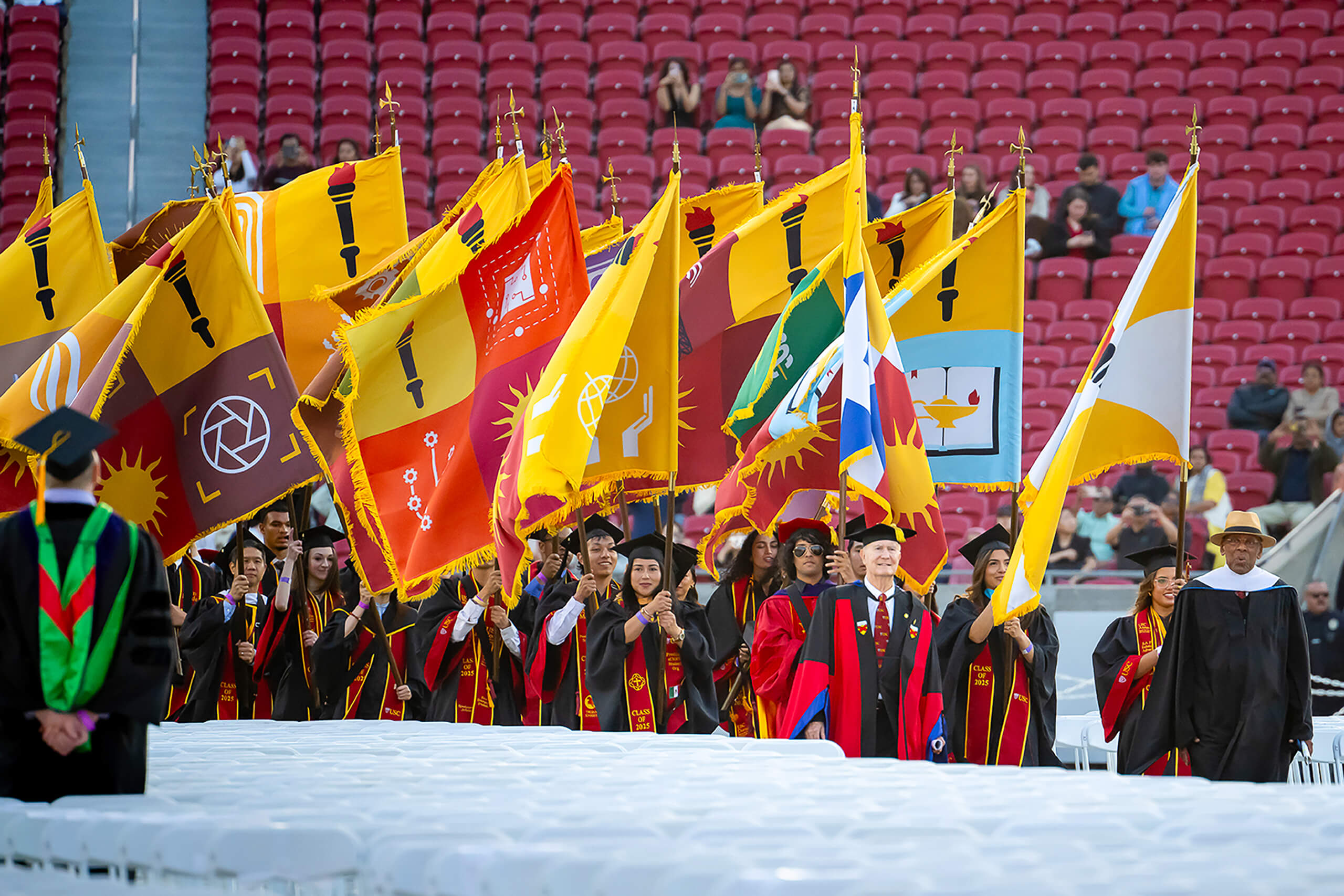 USC graduates in red and gold regalia carry heraldic flags into the Los Angeles Coliseum for the 2025 commencement ceremony.