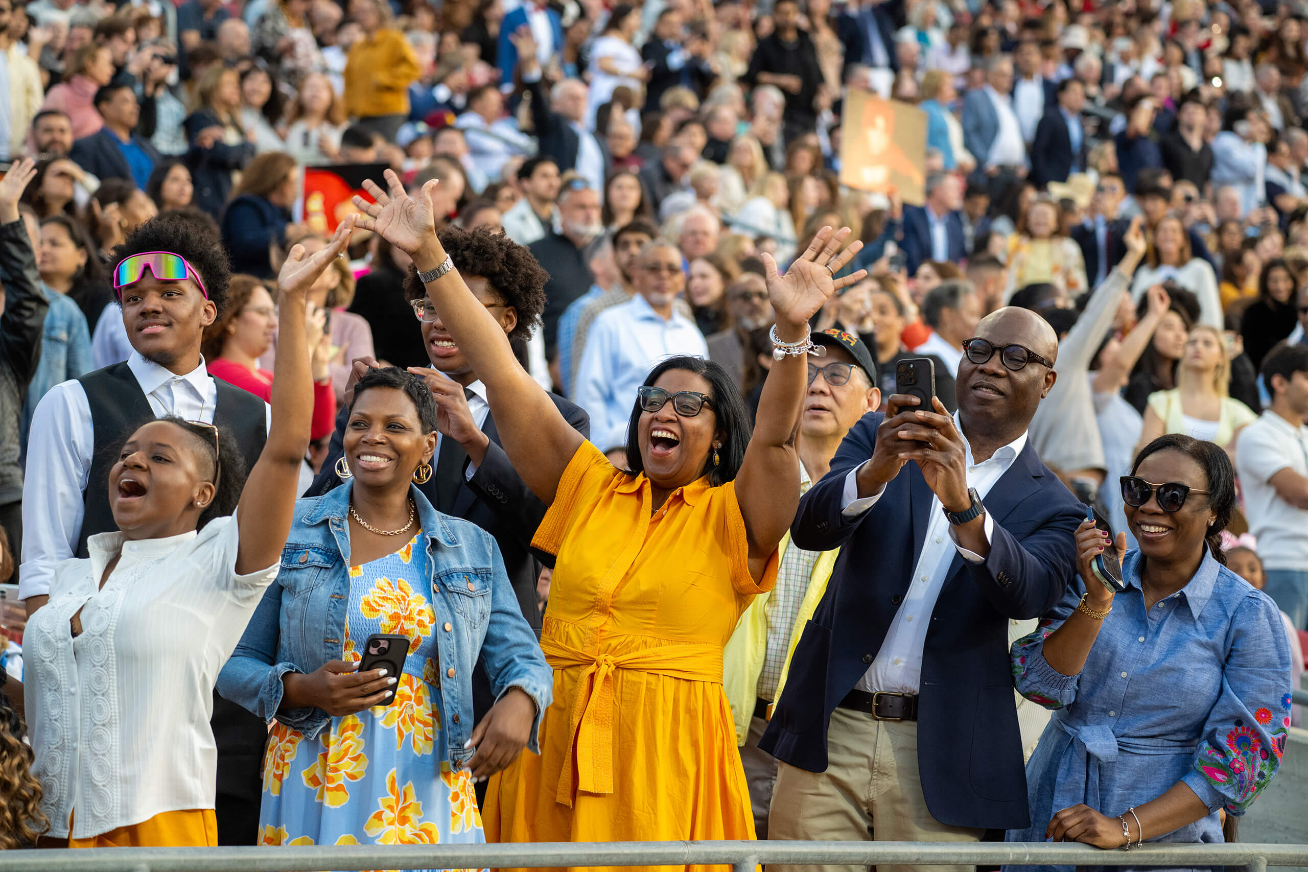 Excited crowd cheers and takes photos at the Los Angeles Coliseum during the 2025 USC commencement ceremony, with hands raised in celebration.