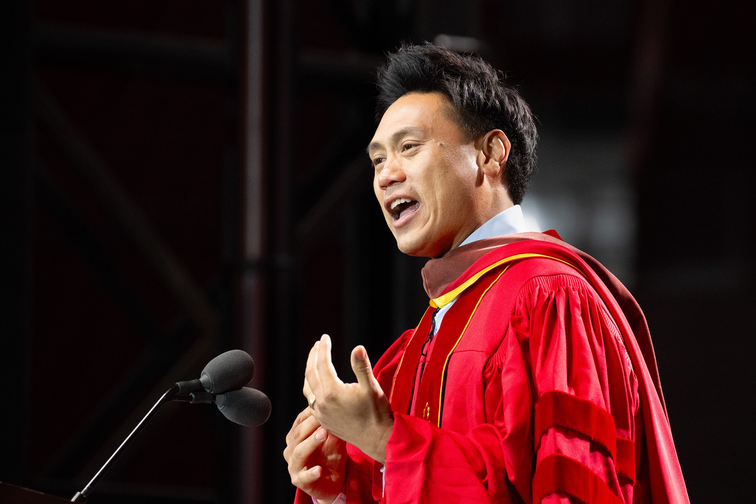 John Chu speaks passionately at a podium, wearing USC red and gold regalia, with microphones and a dark background at the Los Angeles Coliseum ceremony.