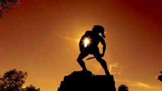 Silhouetted Tommy Trojan statue against a dark orange sunset sky