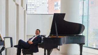 Donald Vega sitting on a bench leaning on a piano in practice room at The Juilliard School in NYC