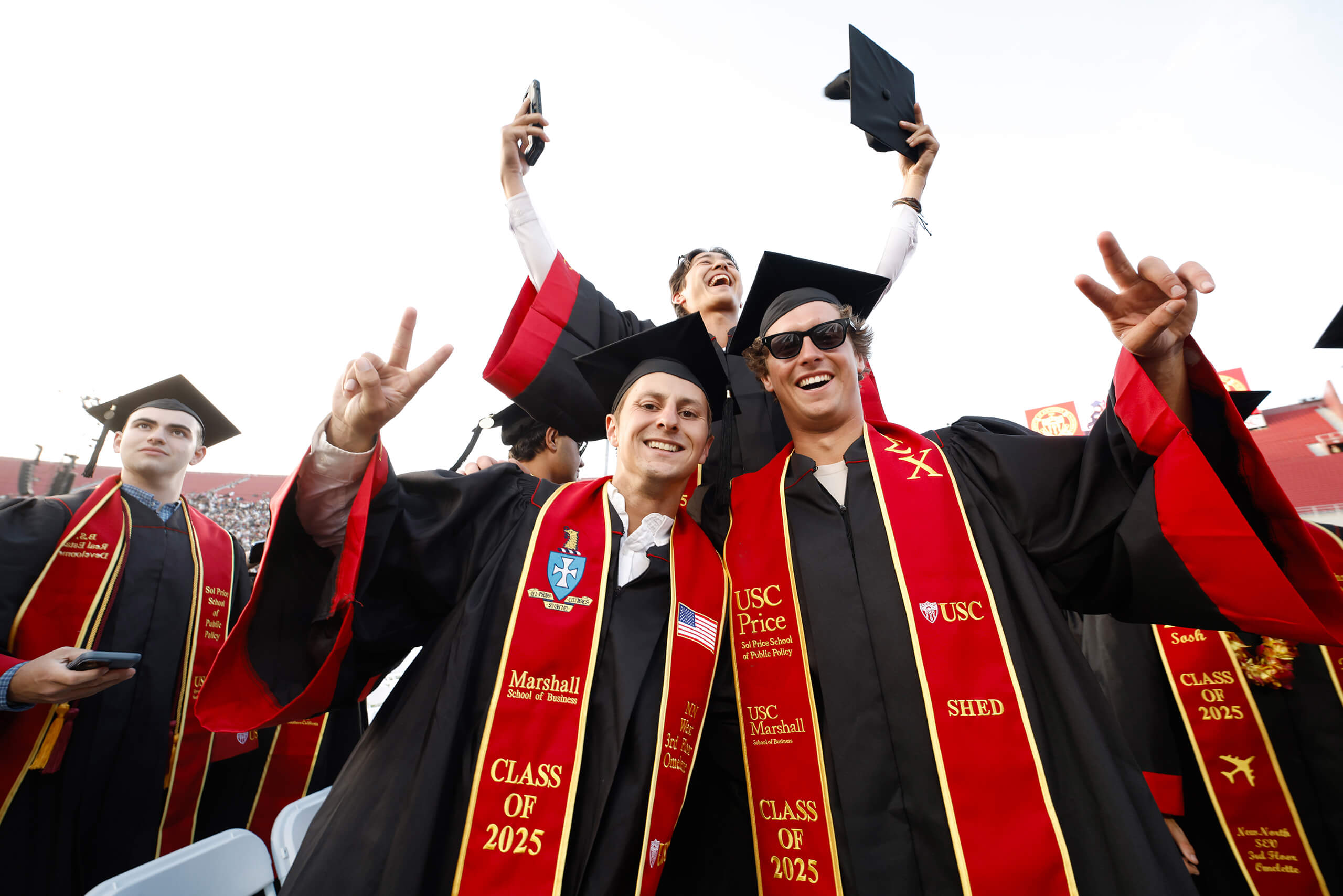 Joyful USC graduates in red and gold regalia from the Class of 2025 make fight on gestures at the Los Angeles Coliseum commencement.