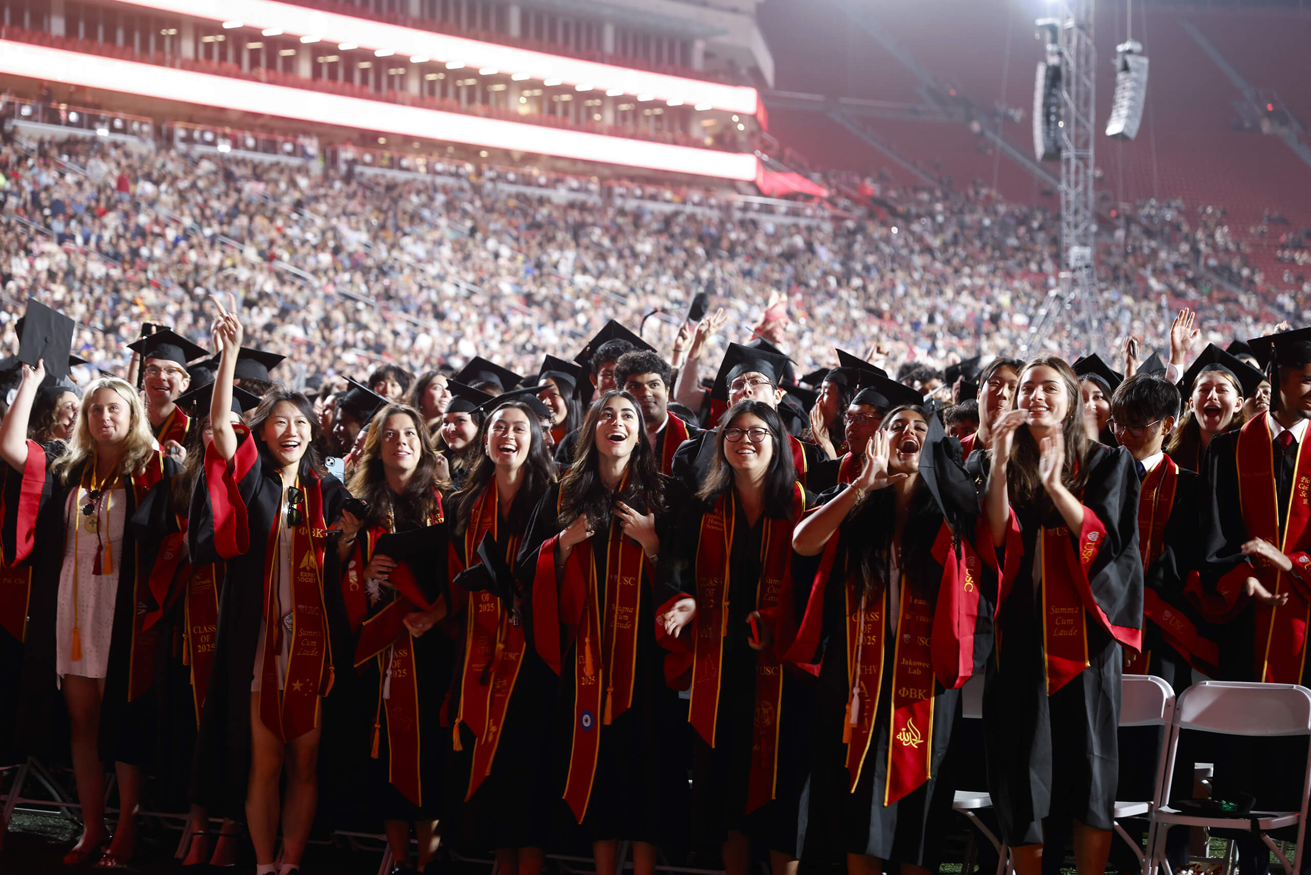 USC graduates in red and gold regalia from the Class of 2025 smile and make the fight on hand gesture while entering the Los Angeles Coliseum for commencement.