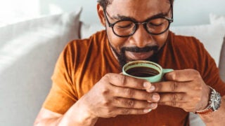 A man wearing black glasses, a beard and a brown shirt sipping a mint green cup filled with dark brown coffee with white pillows in the background