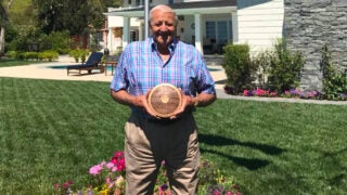 Rink Babka, in a blue plaid shirt and khaki pants holding a brown disk with pink flowers, green grass and a white house in the background