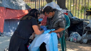 Two women in black clothing and blue gloves helping a man wearing a gray jacket and sitting in a green chair with a tent in the background