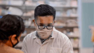 A pharmacist wearing a white mask, white robe, and black glasses shows an orange prescription bottle to a customer.