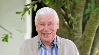 B. Wayne Hughes smiling and wearing a tan jacket and a striped light blue shirt in front of a green, black and white background
