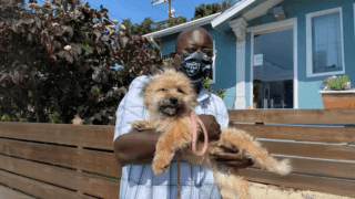 Charles, wearing a black Raiders mask and holding a light brown dog in front of a light blue house
