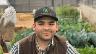 Jose Ruiz, wearing a black cap, dark brown vest and a black and white plaid shirt, holding a shovel in front of a community garden