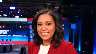 Rachel Scott, wearing a red suit and white shirt, sitting and smiling in front of a blue and red newsroom