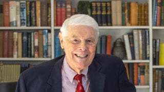 Tom Capehart, smiling and wearing a navy suit jacket, red and white striped shirt and a red and gold striped tie, with a white bookcase full of books in the background