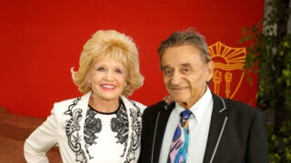 From left, Charlotte Ginsburg wearing a black and white suit and Allen Ginsburg wearing a black suit jacket, white shirt and colorful tie, posing in front of a cardinal wall with a gold shield on it