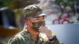 Lt. Justin Lee holds his hand up to his face while wearing a camouflage hat, face mask and uniform, clear gloves and a black watch